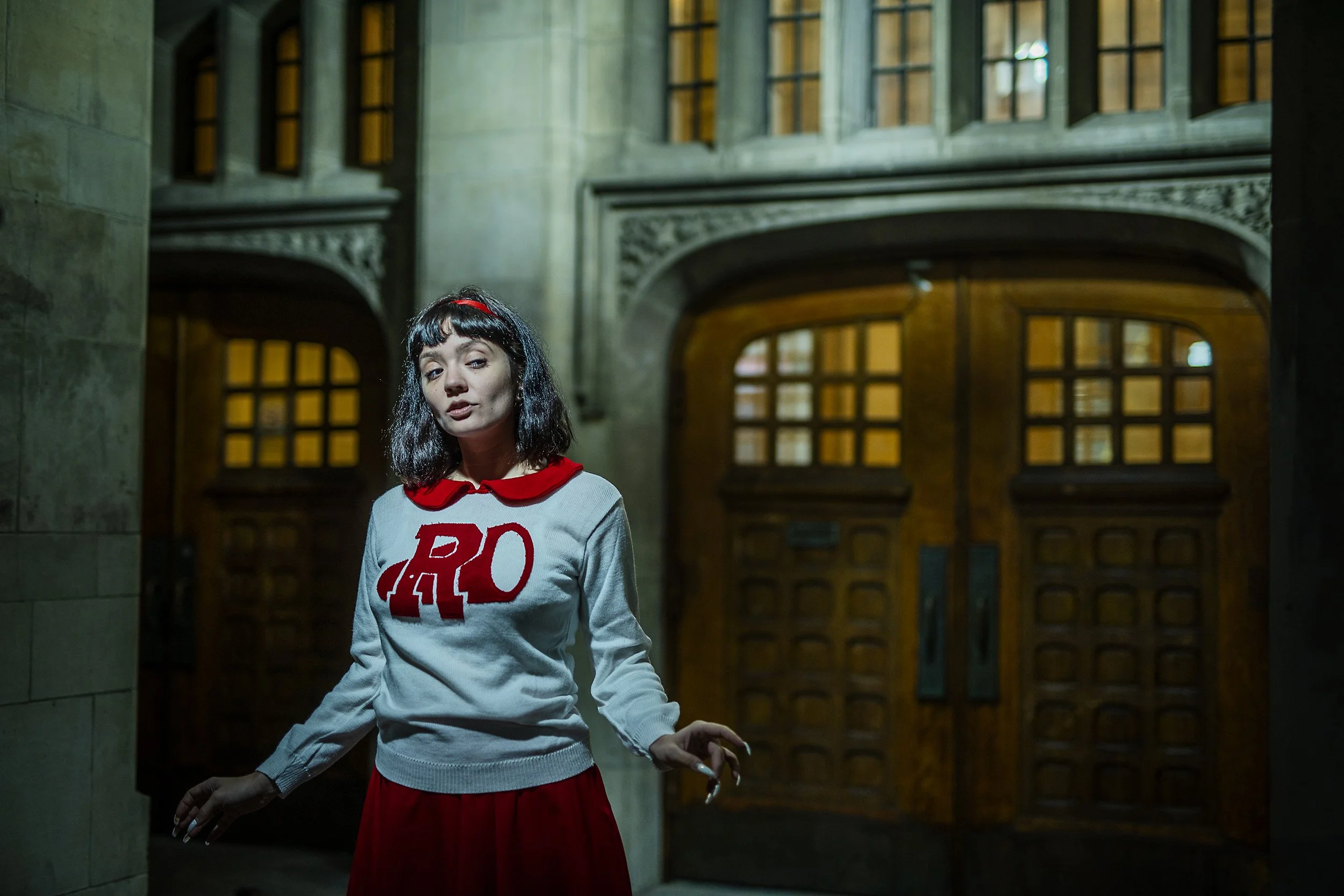 A young woman with black hair and a red headband posing indoors. She is wearing a gray sweater with large red letters and red trim, and a red skirt. The background features a large wooden door with glass panes and stone walls.