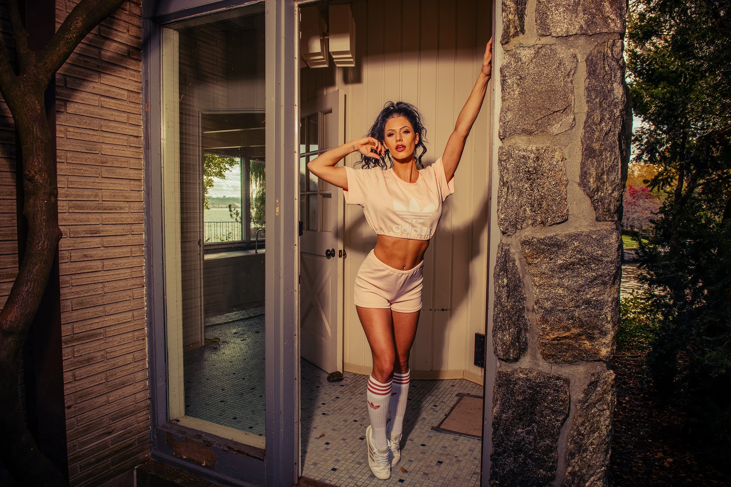 A woman stands in an entryway of a house, posing with one arm raised and the other hand near her face. She is wearing a light pink Adidas crop top, shorts, knee-high socks, and sneakers. The scene is taken during sunset, and the house features a bric
