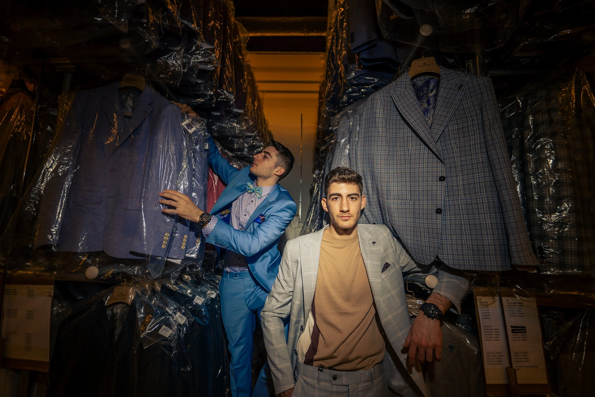 Two men shopping for suits in a store aisle filled with wrapped clothing and boxes, one in a light gray plaid suit and the other in a bright blue suit, with stacked boxes visible on the lower shelves.