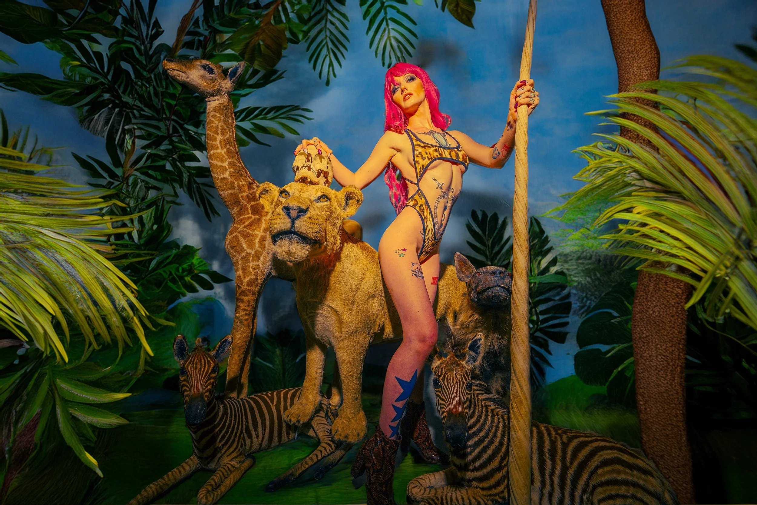 A woman with pink hair and tattoos posing with a giraffe, lion, tiger, and zebra in a jungle-themed setting with green leaves and trees.
