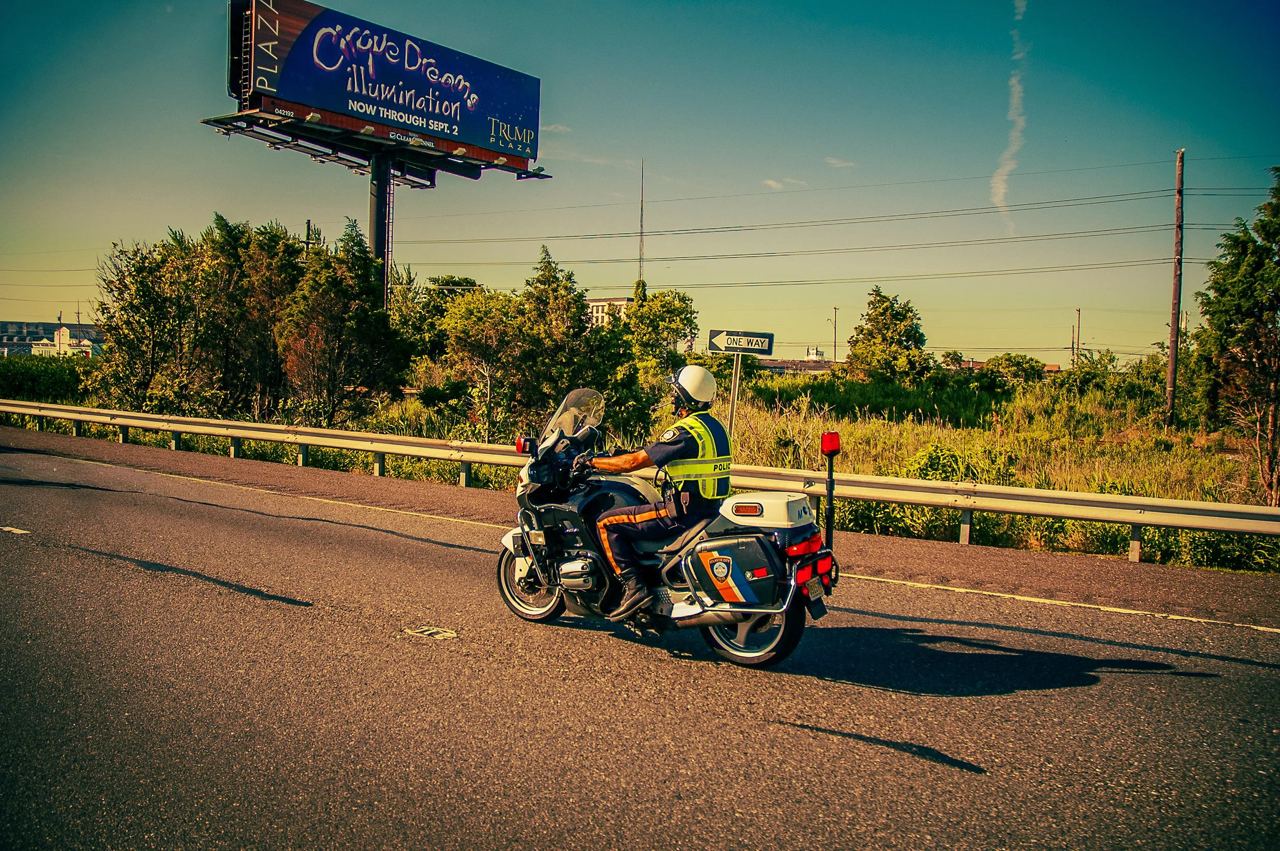 A police officer on a motorcycle on the side of a highway, with a billboard advertising Cirque Dreams Illumination and a one-way traffic sign in the background
