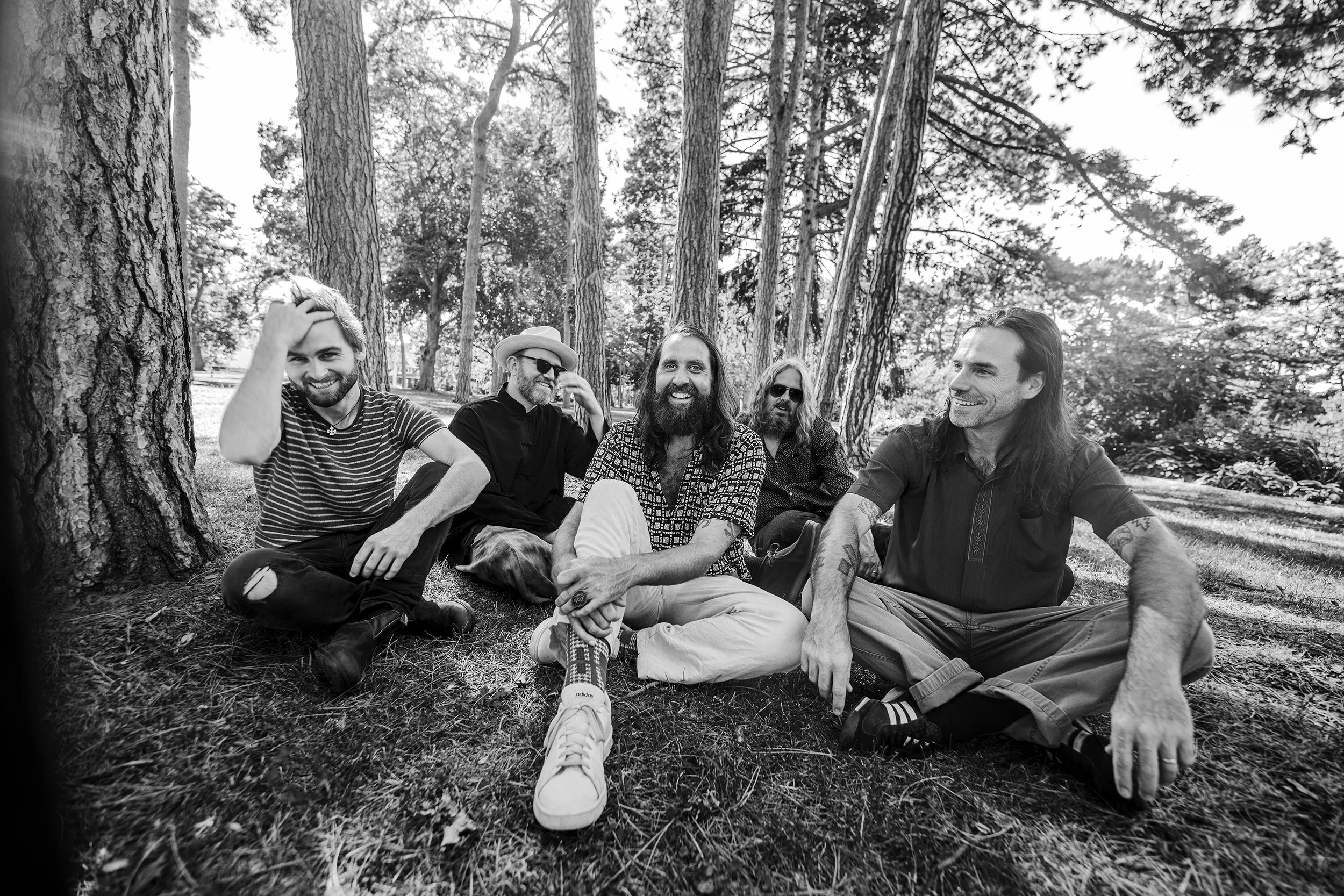 A group of five men sitting on grass in a wooded park, smiling and relaxing together.