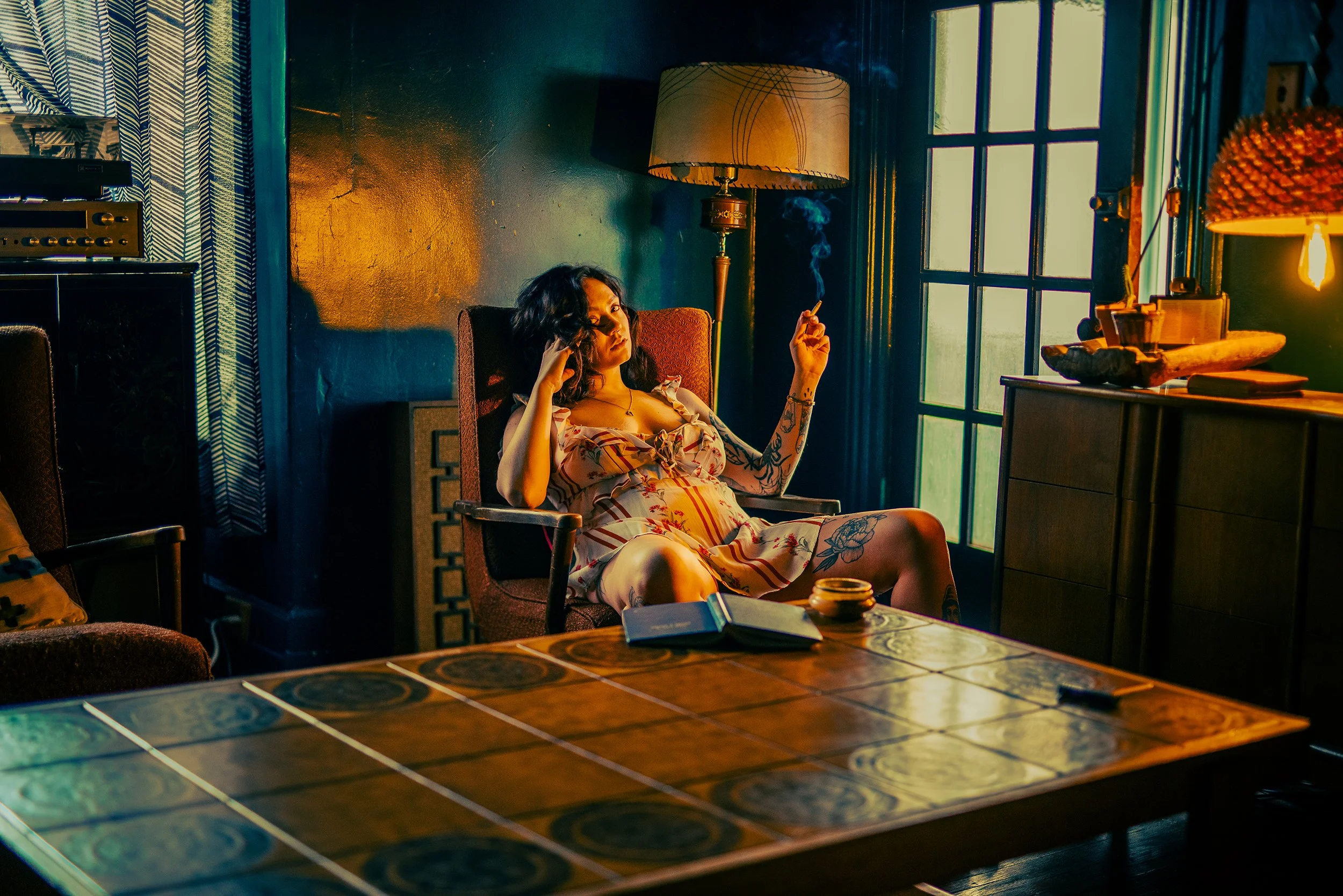 A woman sitting comfortably in a mid-century modern armchair in a dimly lit room, smoking a cigarette, with a relaxed expression. The room features colorful lighting, a large table with tiled surface in the foreground, and a window with sunlight stre