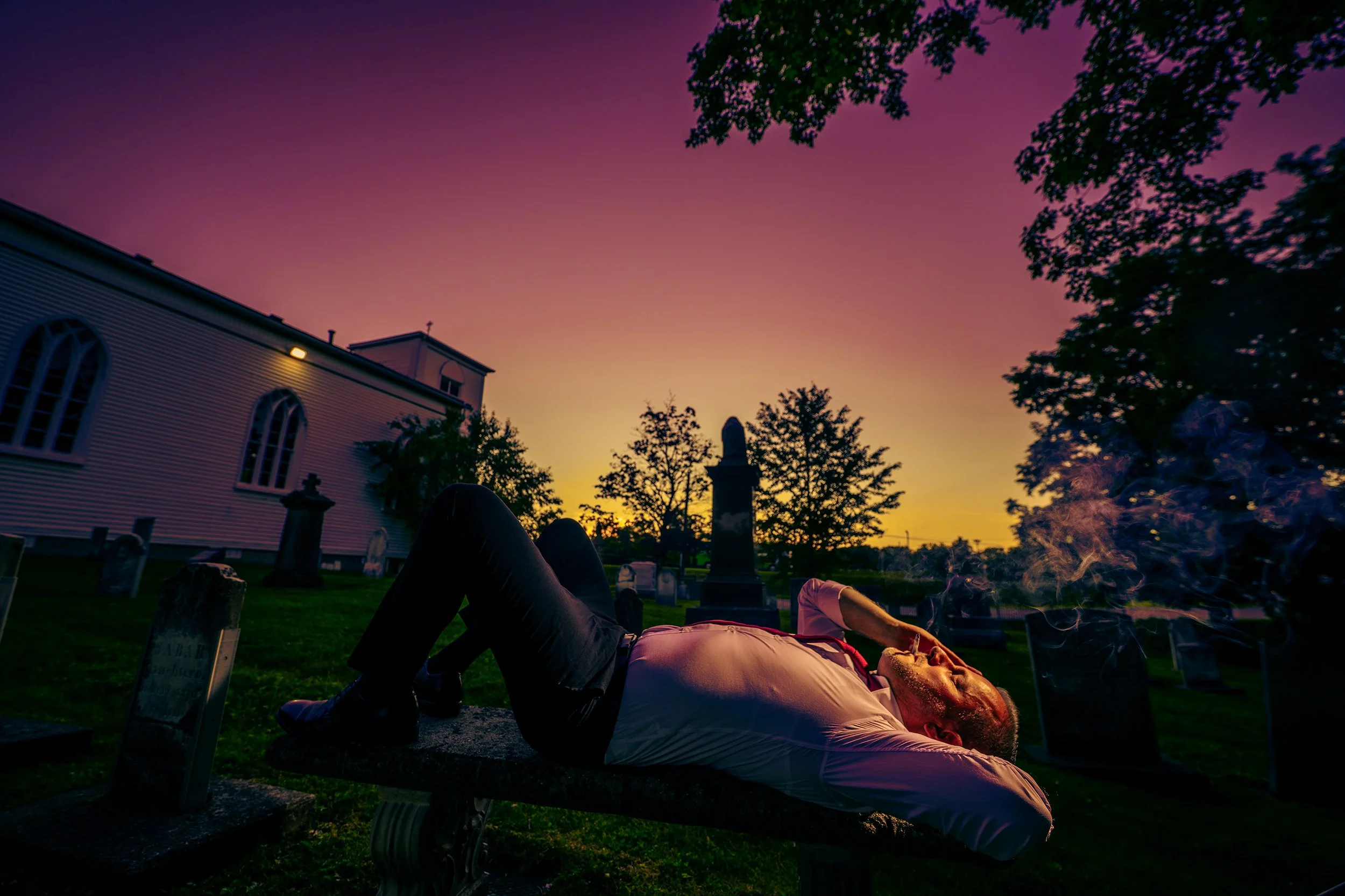 Person lying on a park bench during sunset, with a smoky fire nearby, a church building with arched windows and a steeple, and trees with pink, purple, and orange sky in the background.