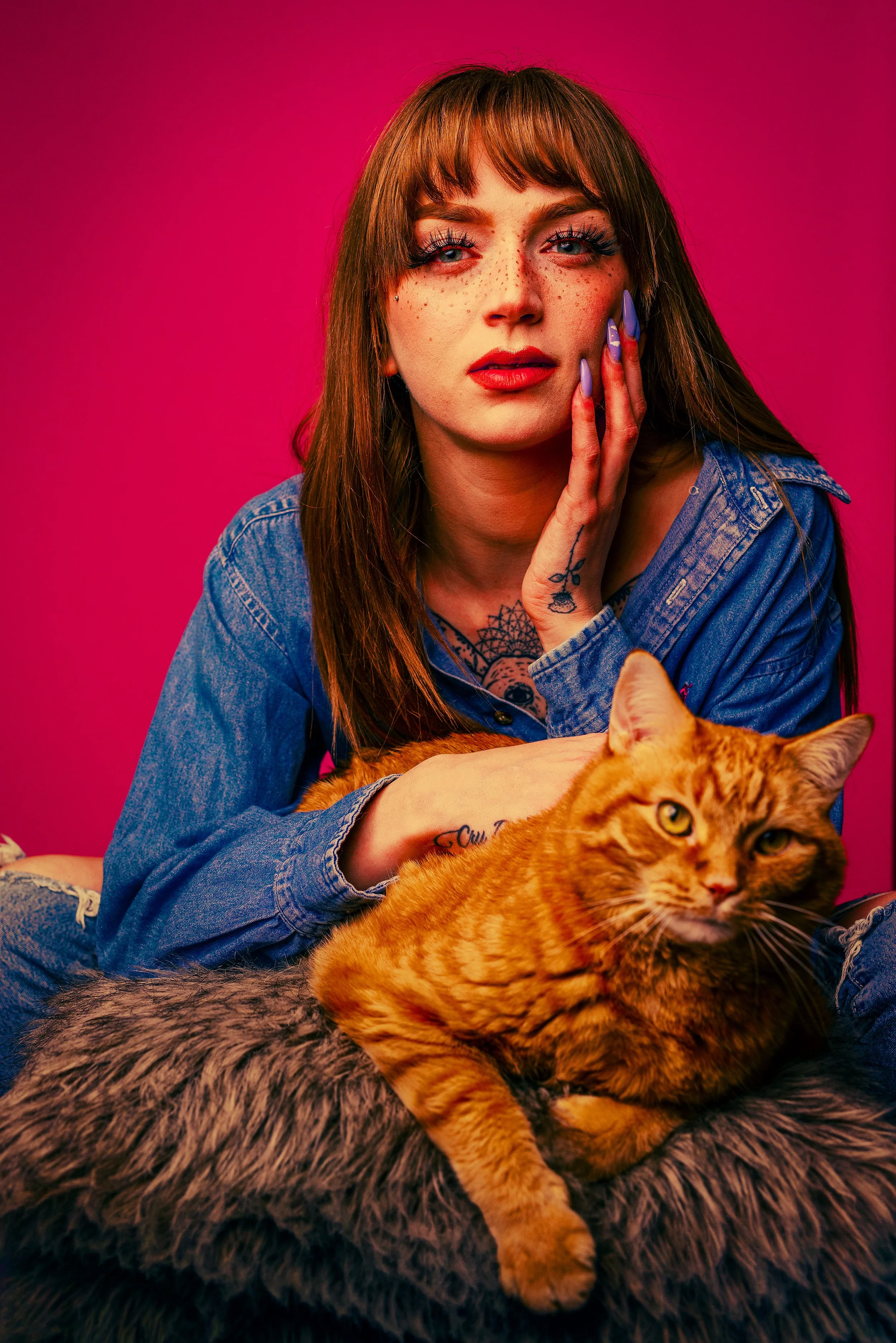 A young woman with red hair, freckles, and blue eyes, wearing a denim jacket, touches her face with one hand, and holds an orange tabby cat on her lap. The background is a vibrant pink.