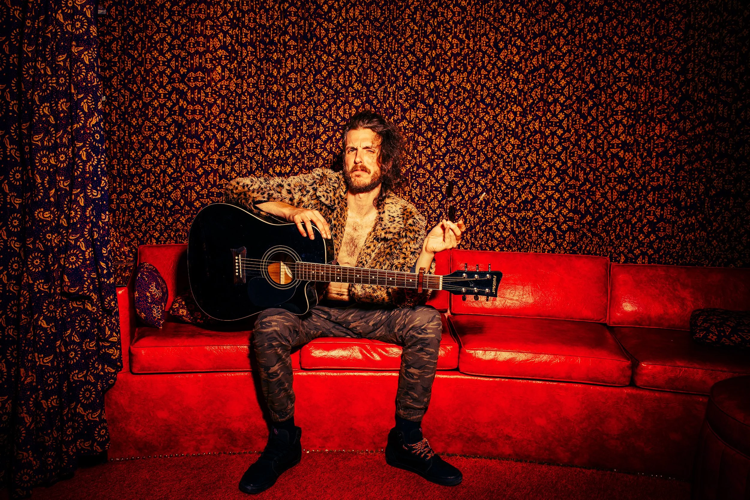 A man with long hair and a beard sitting on a red sofa, holding an acoustic guitar, against a patterned dark background with drapes on the left.