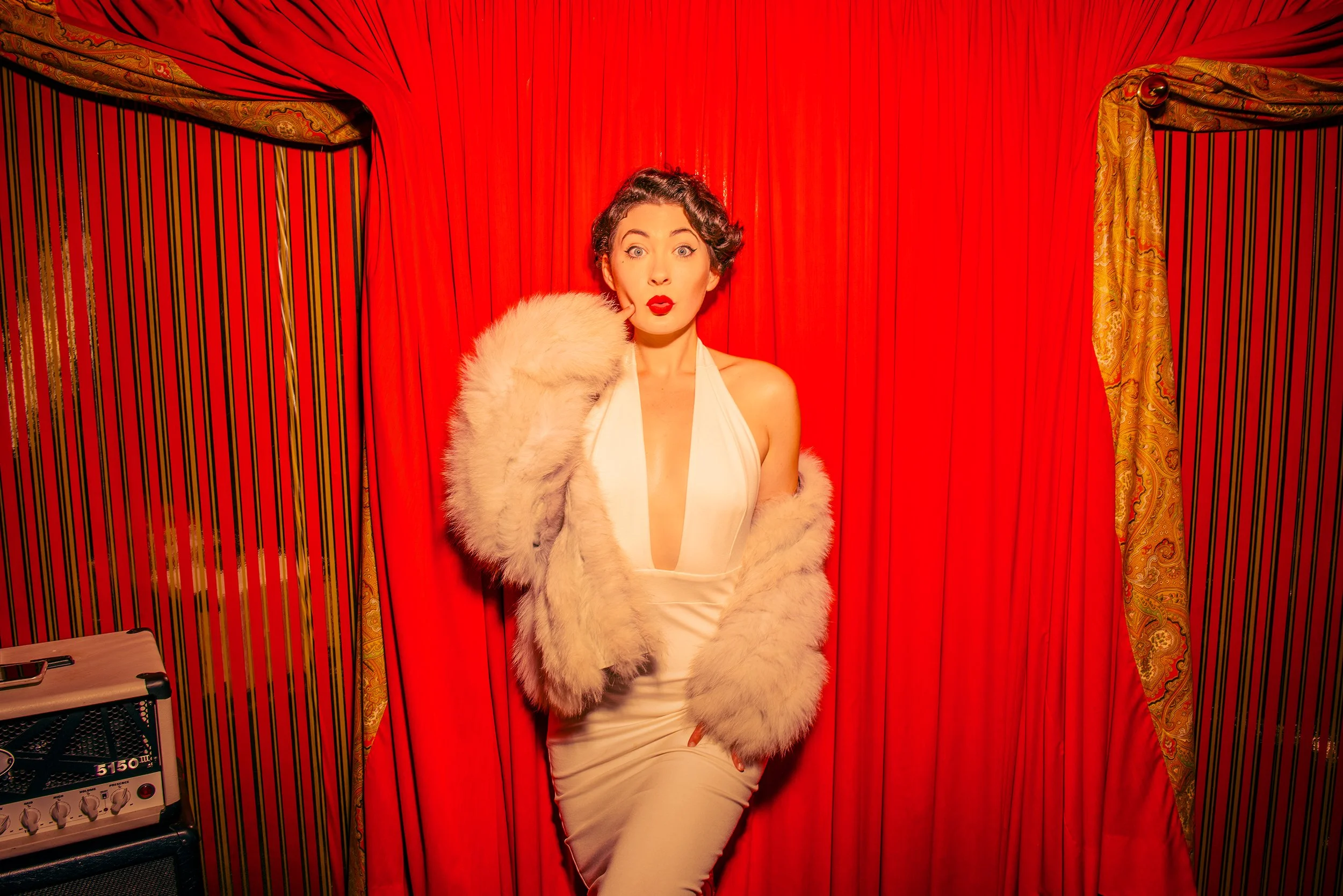 A woman dressed in a vintage style with red lipstick, white dress, and faux fur coat, standing in front of red curtains with gold accents.