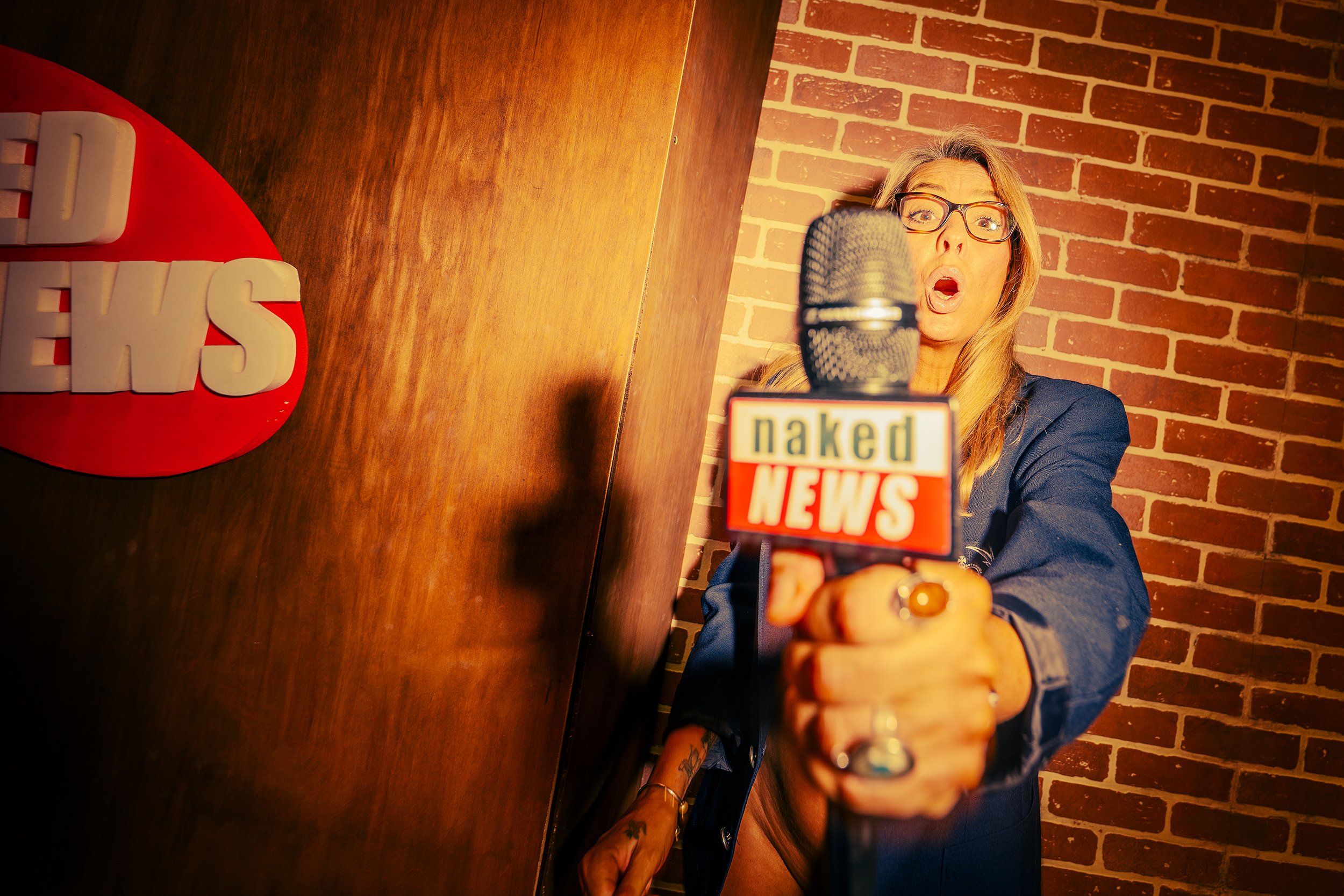 A woman with glasses holding a microphone with a "naked news" logo, standing in front of a brick wall, with part of a large red sign that says "NEWS" visible on the desk.