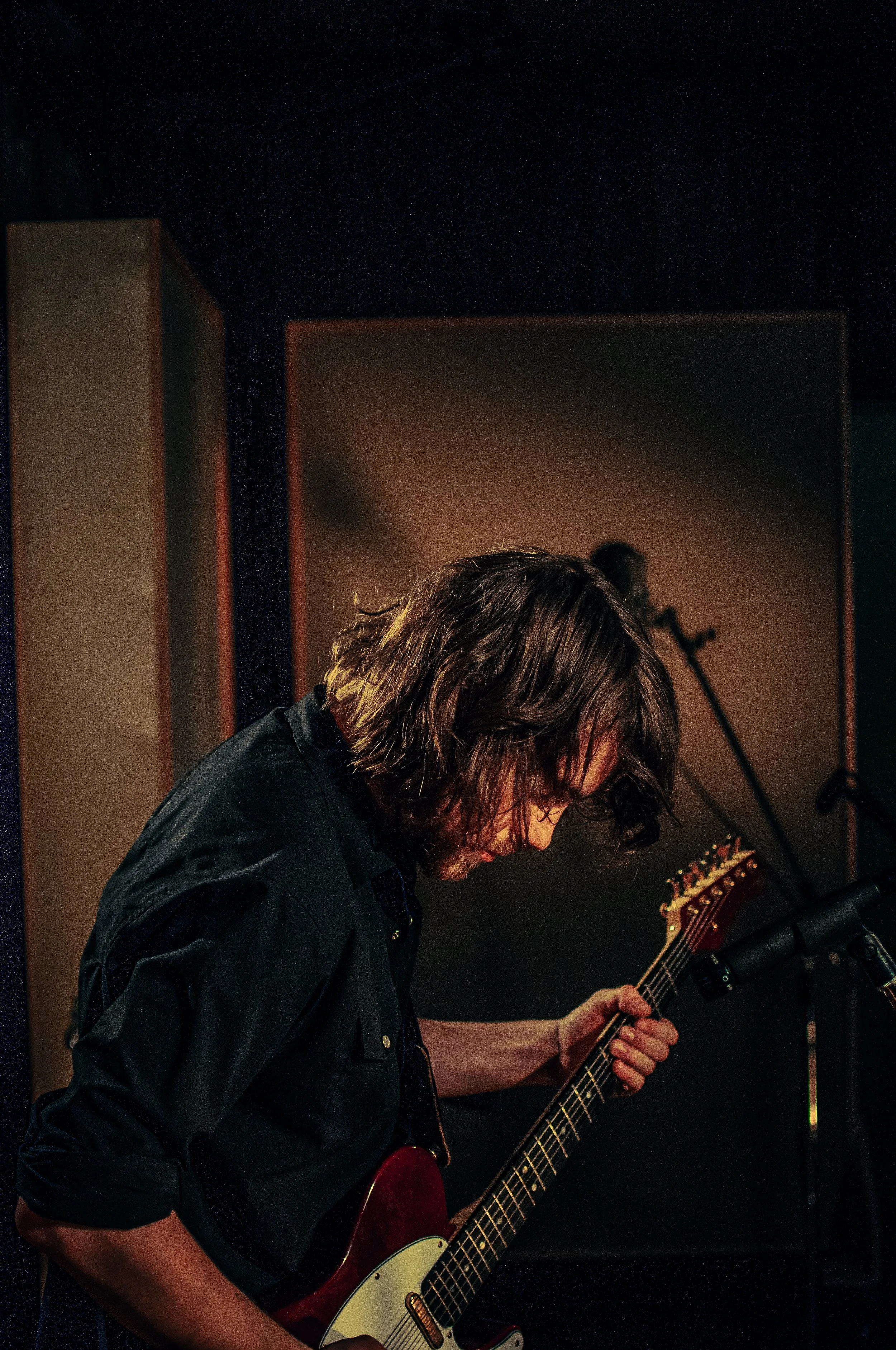 A man with shoulder-length dark hair and glasses playing an electric guitar on stage, with a microphone and a music stand nearby, in a dimly lit setting.
