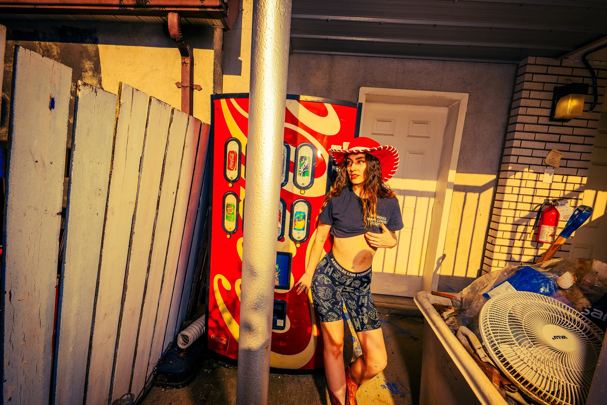 A young woman wearing a large red sombrero, a navy blue t-shirt, and patterned shorts stands next to a Coca-Cola vending machine outside a building with a brick wall and a white door. The woman is looking to the side with her hand on her hip, and the