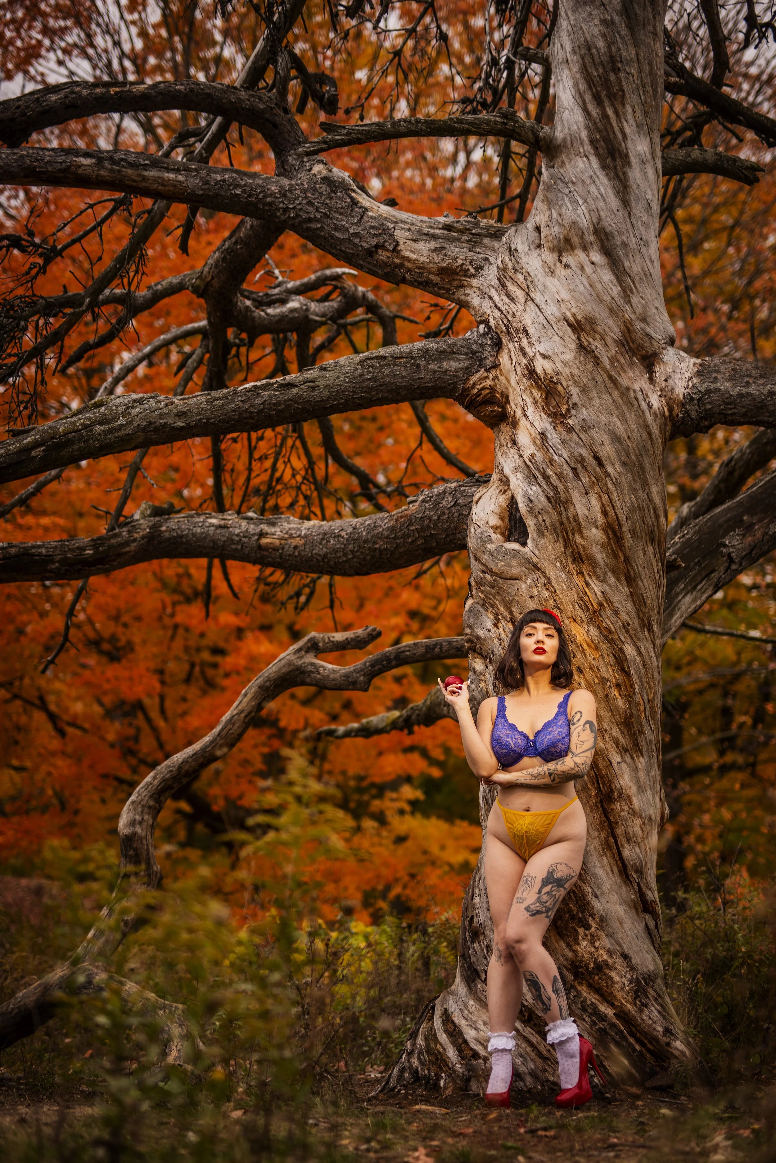 A woman with tattoos standing next to a large gnarled tree with autumn leaves in the background, holding an apple in her right hand.