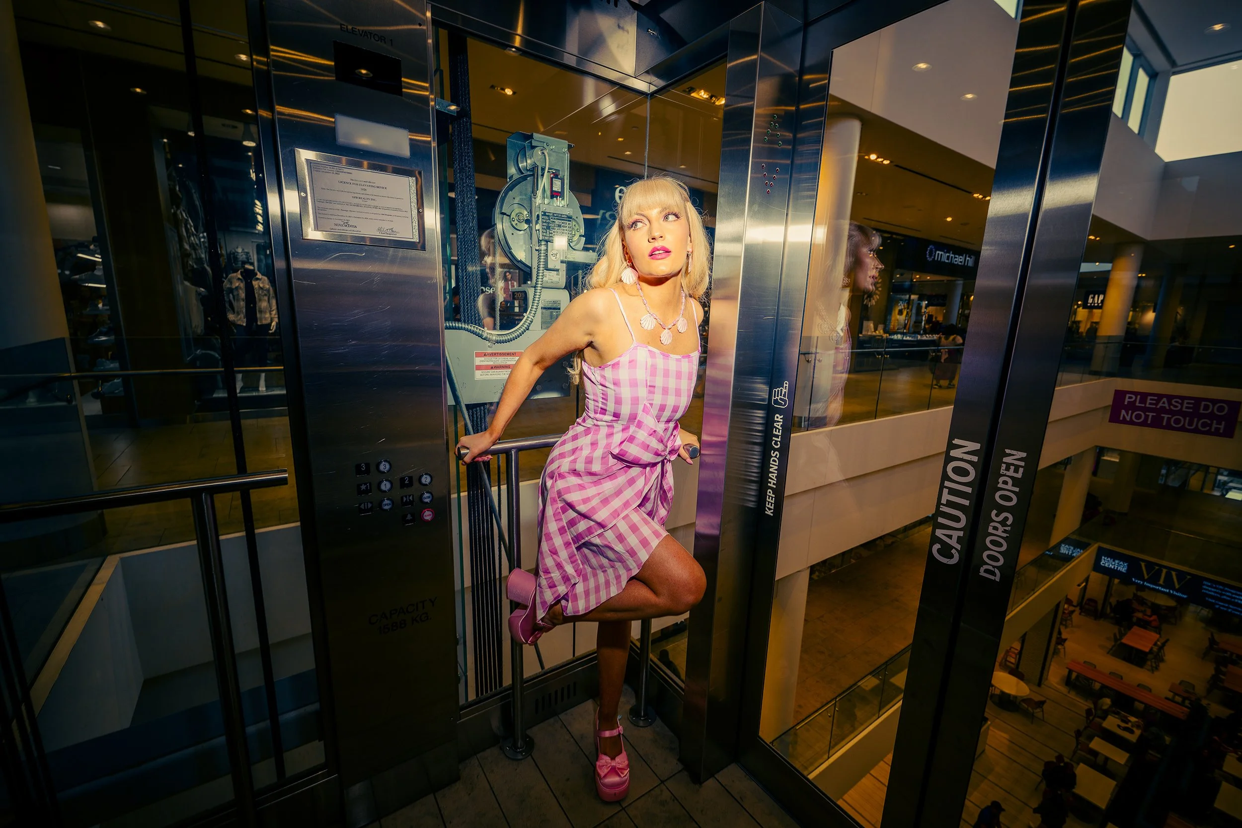 A woman standing in an elevator shaft in a shopping mall, wearing a pink and white checkered dress, pink shoes, and jewelry, with one leg raised and her hand on the railing, looking away from the camera.