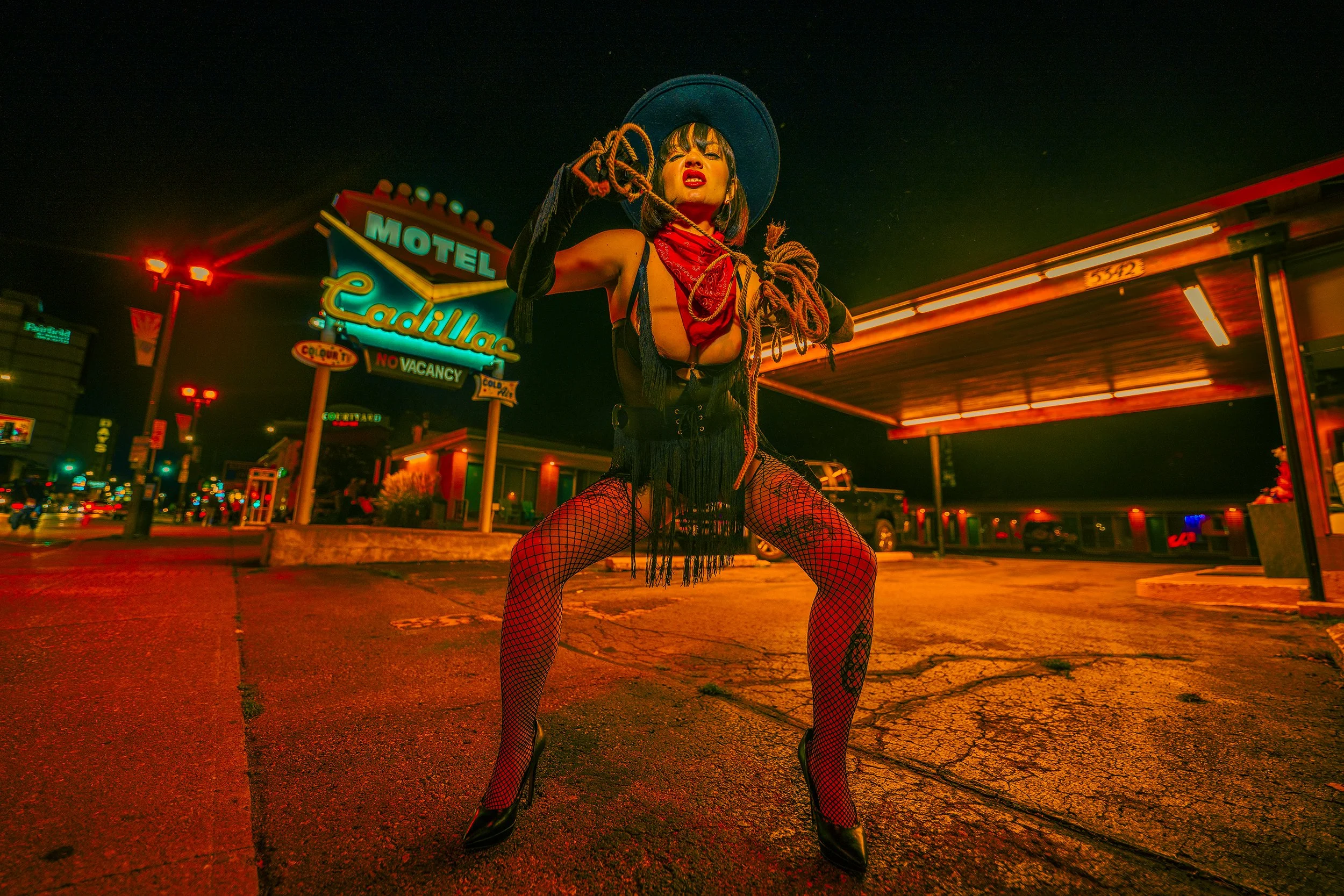 A person dressed in a cowgirl outfit with fishnet stockings, high heels, and holding a rope, standing in a neon-lit parking lot at night near a motel called Cadillac.