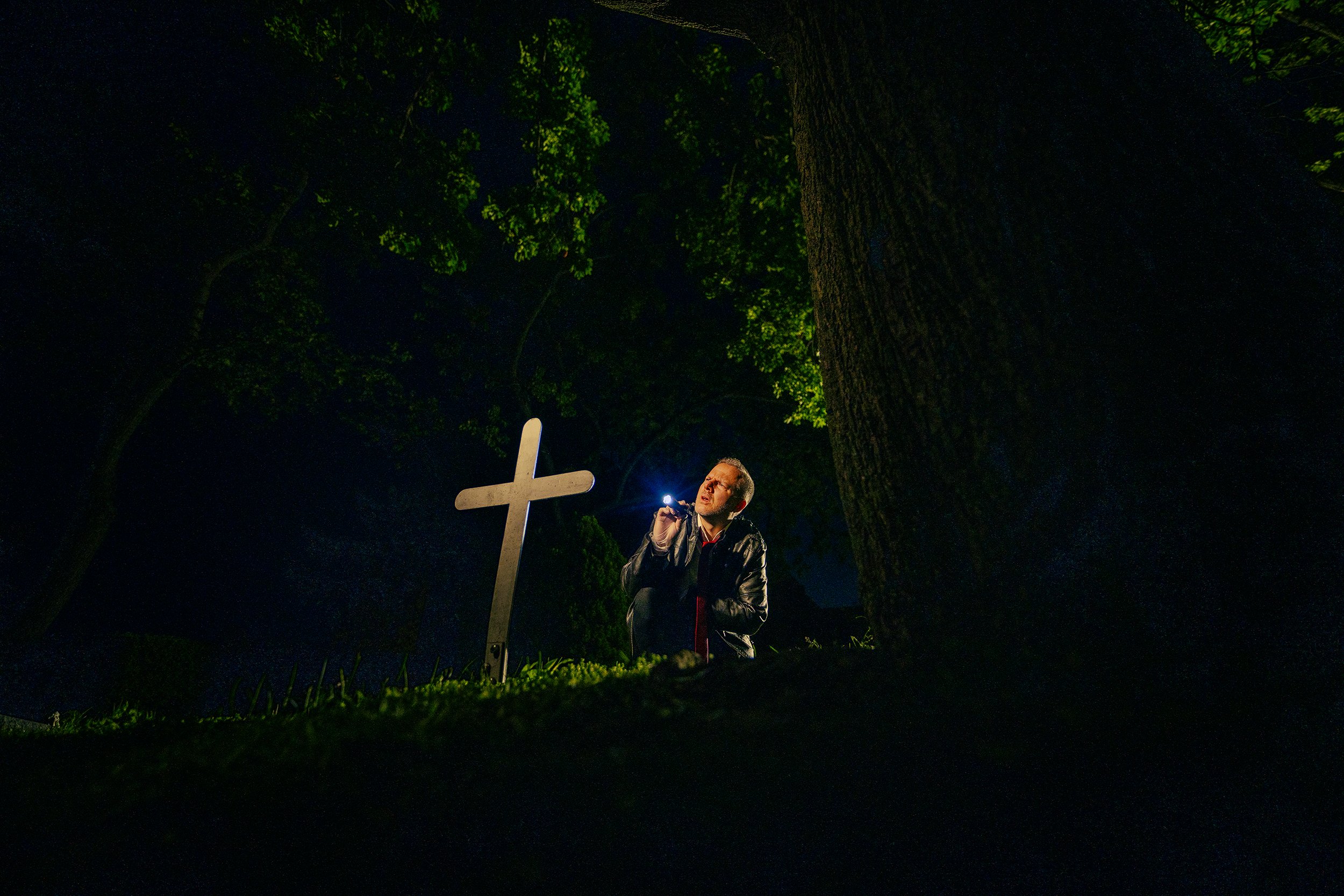 A man with short hair and glasses holding a flashlight in a dark outdoor area with trees. There is a white cross in front of him, and he is looking up towards the sky.