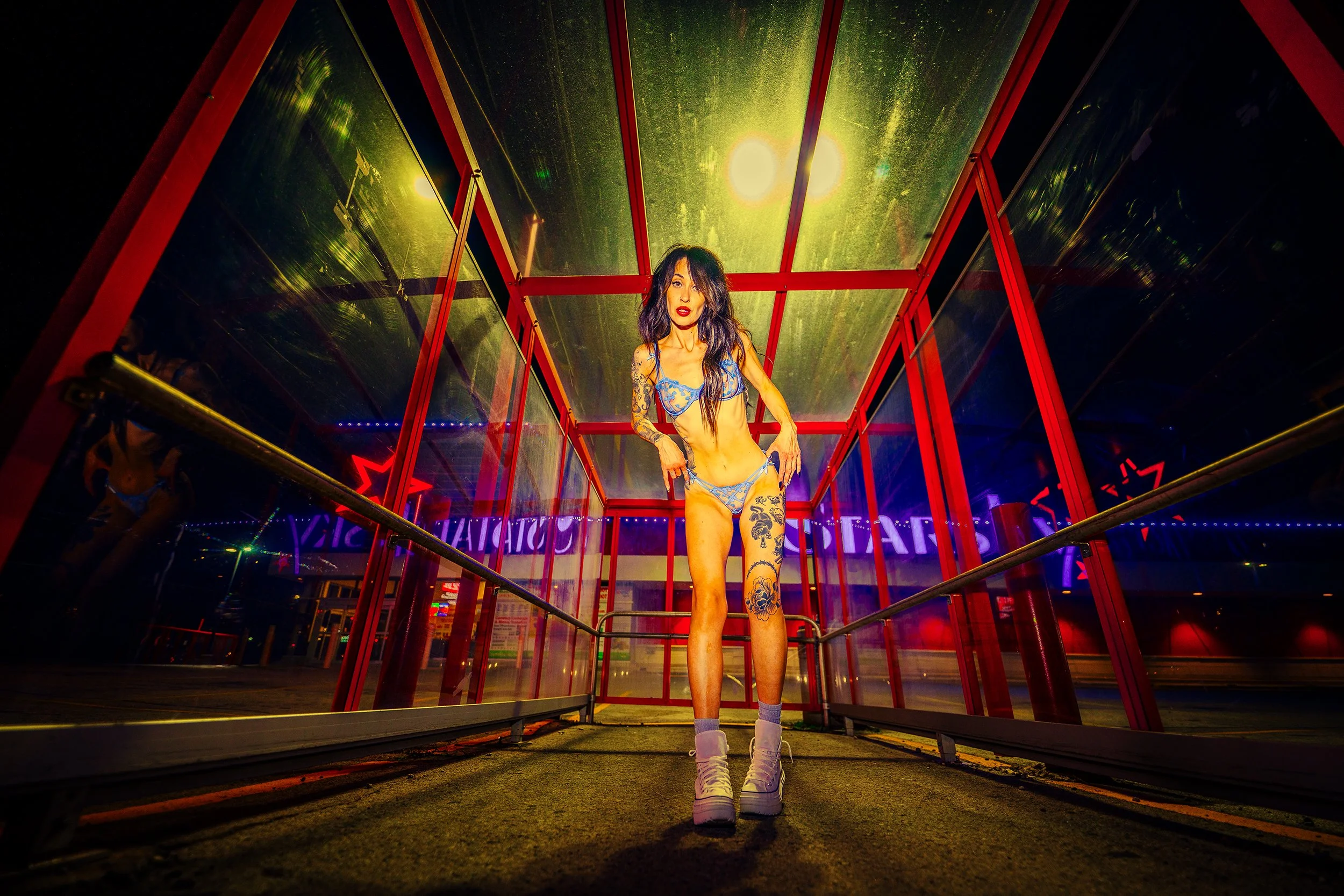 A woman stands inside a glass structure illuminated by yellow and red lights at night, wearing lingerie, boots, and sporting tattoos.