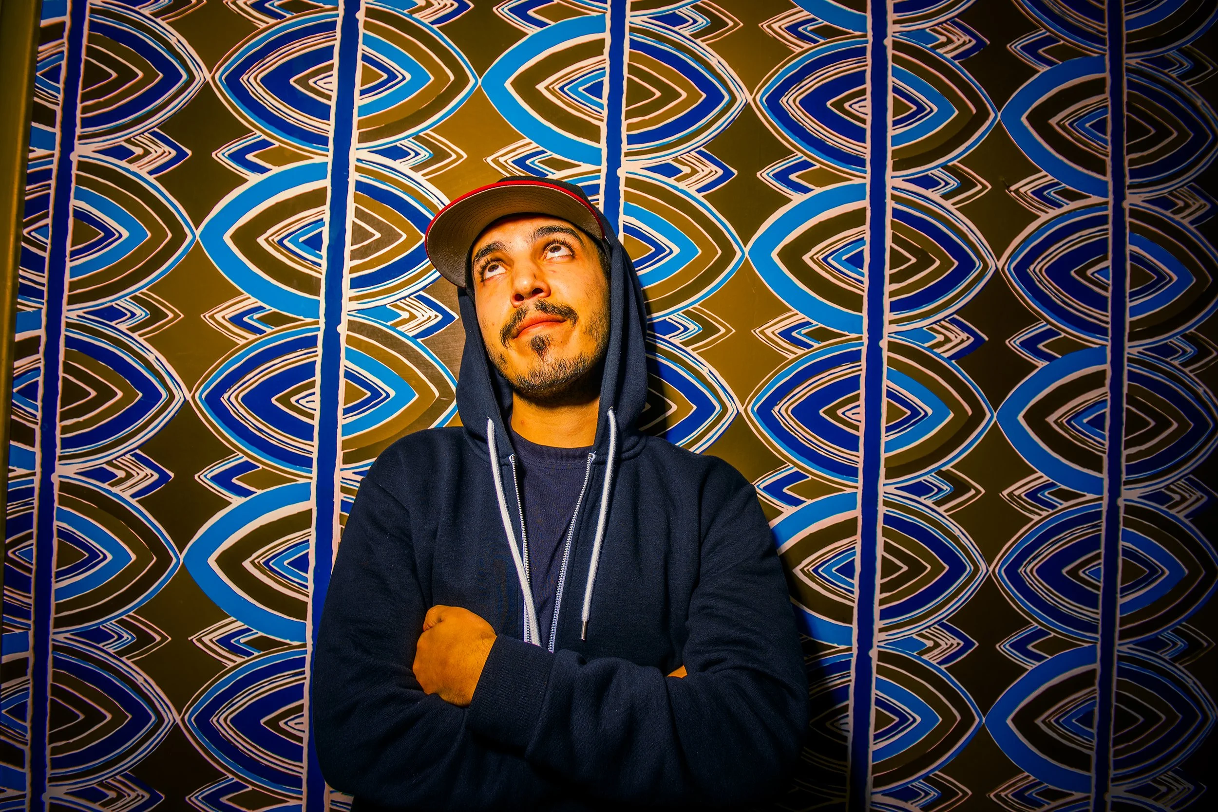 A man wearing a dark hoodie and a baseball cap facing slightly upward with arms crossed, standing in front of a colorful geometric patterned wall made of blue, white, and black concentric oval shapes.