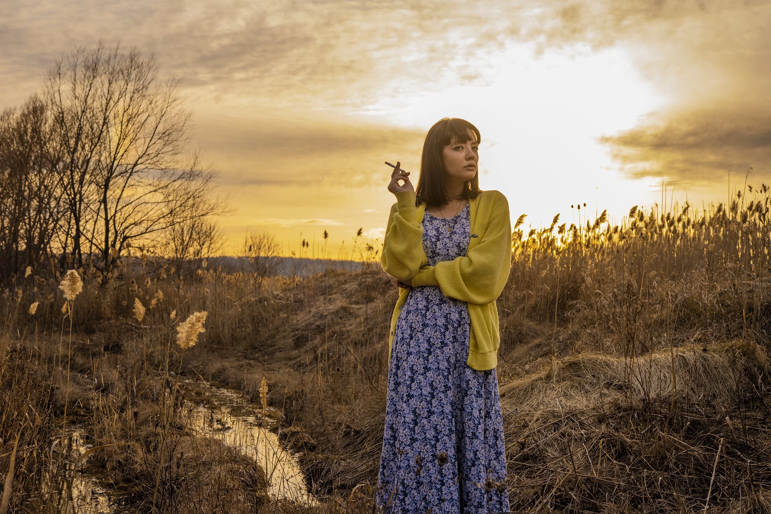 A young woman with shoulder-length dark hair holding a cigarette in her hand, standing outdoors in a field during sunset, wearing a yellow coat over a floral dress, with leafless trees and tall grasses in the background.