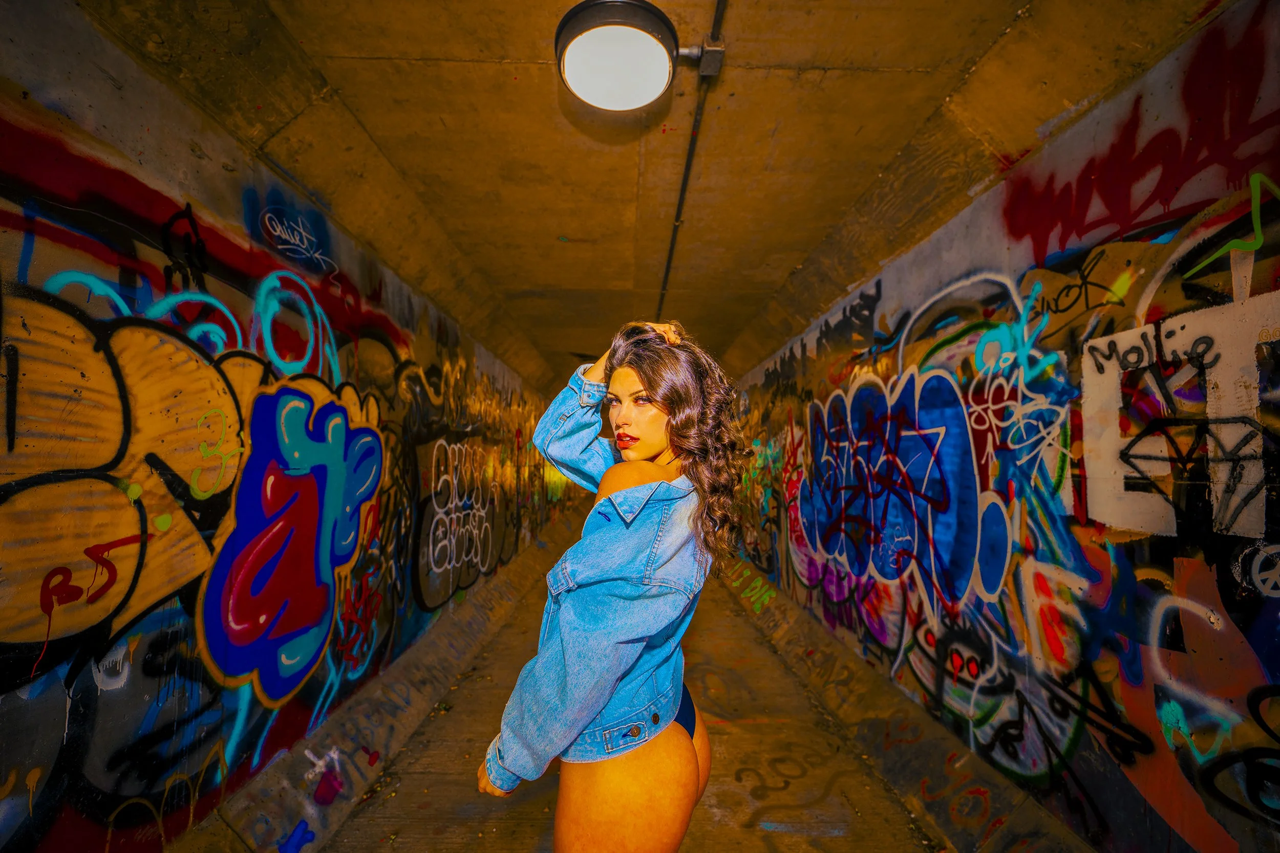 A woman with wavy brown hair poses in a graffiti-covered tunnel, wearing a blue denim jacket and showing her bare lower back.