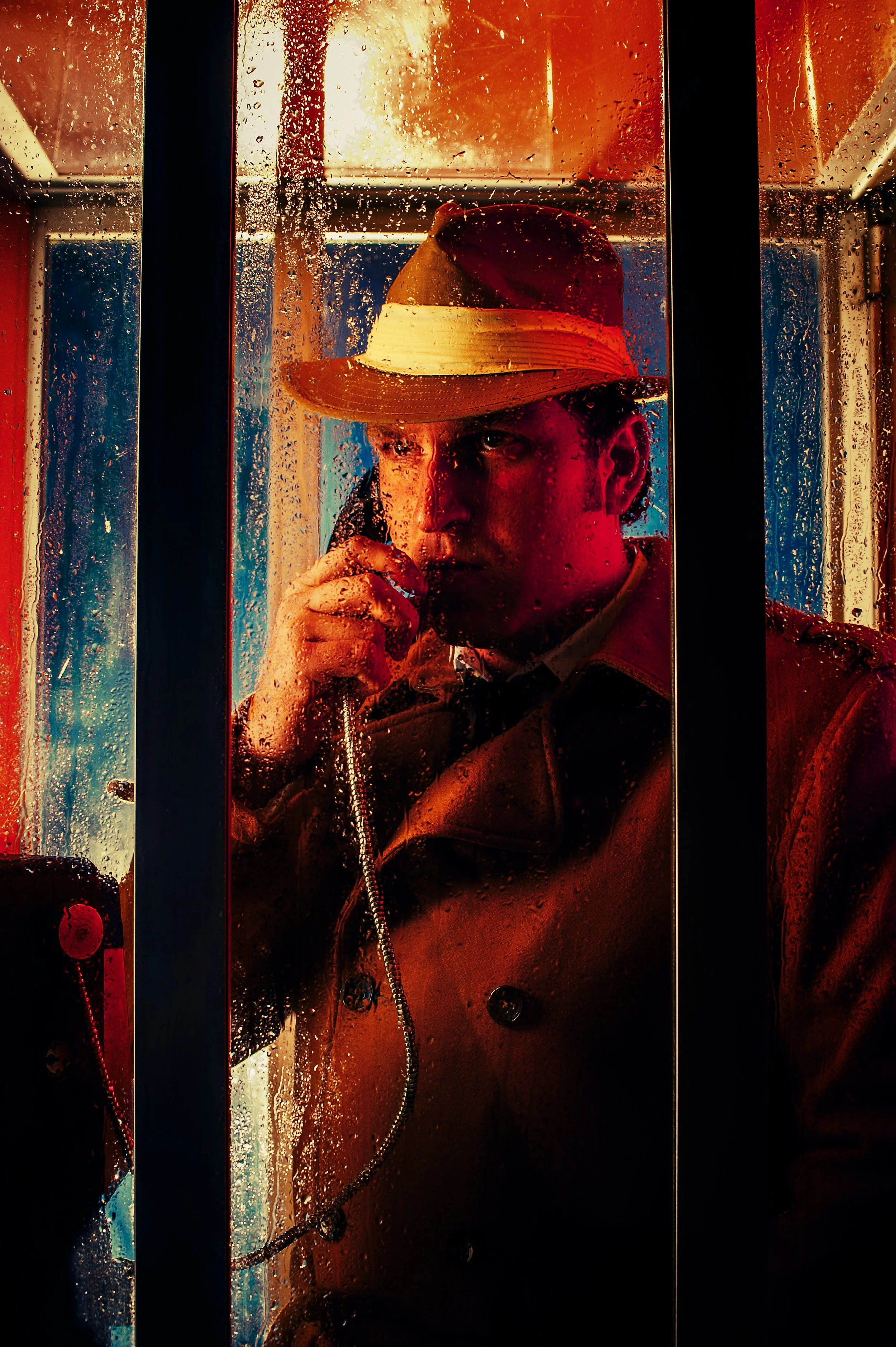 A man in a trench coat and fedora hat talking on a pay phone inside a rain-covered phone booth at night.