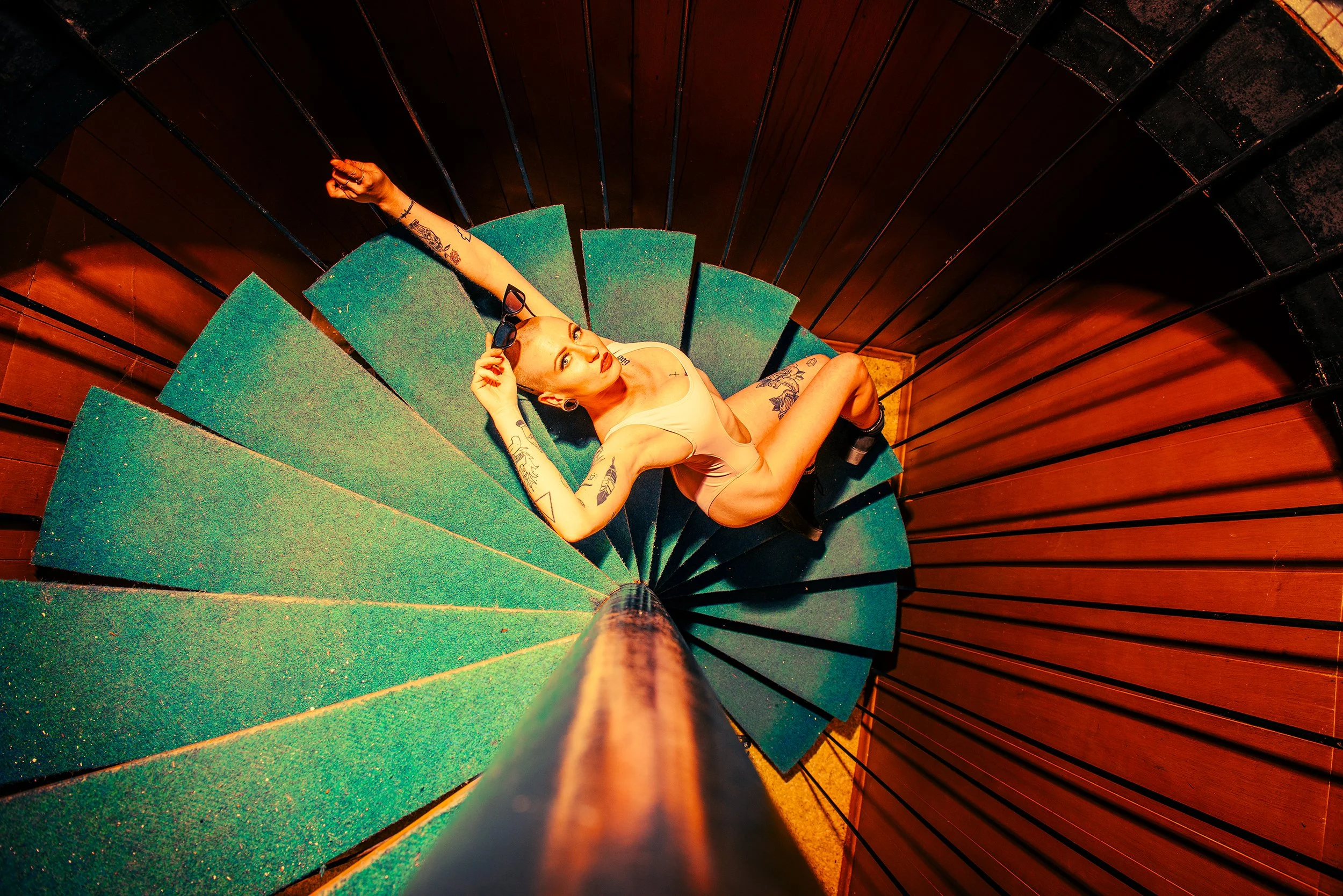 A woman sitting on a green carpeted staircase at the top of a spiral staircase, holding sunglasses in her right hand, with tattoos on her arms and chest, looking up at the camera.