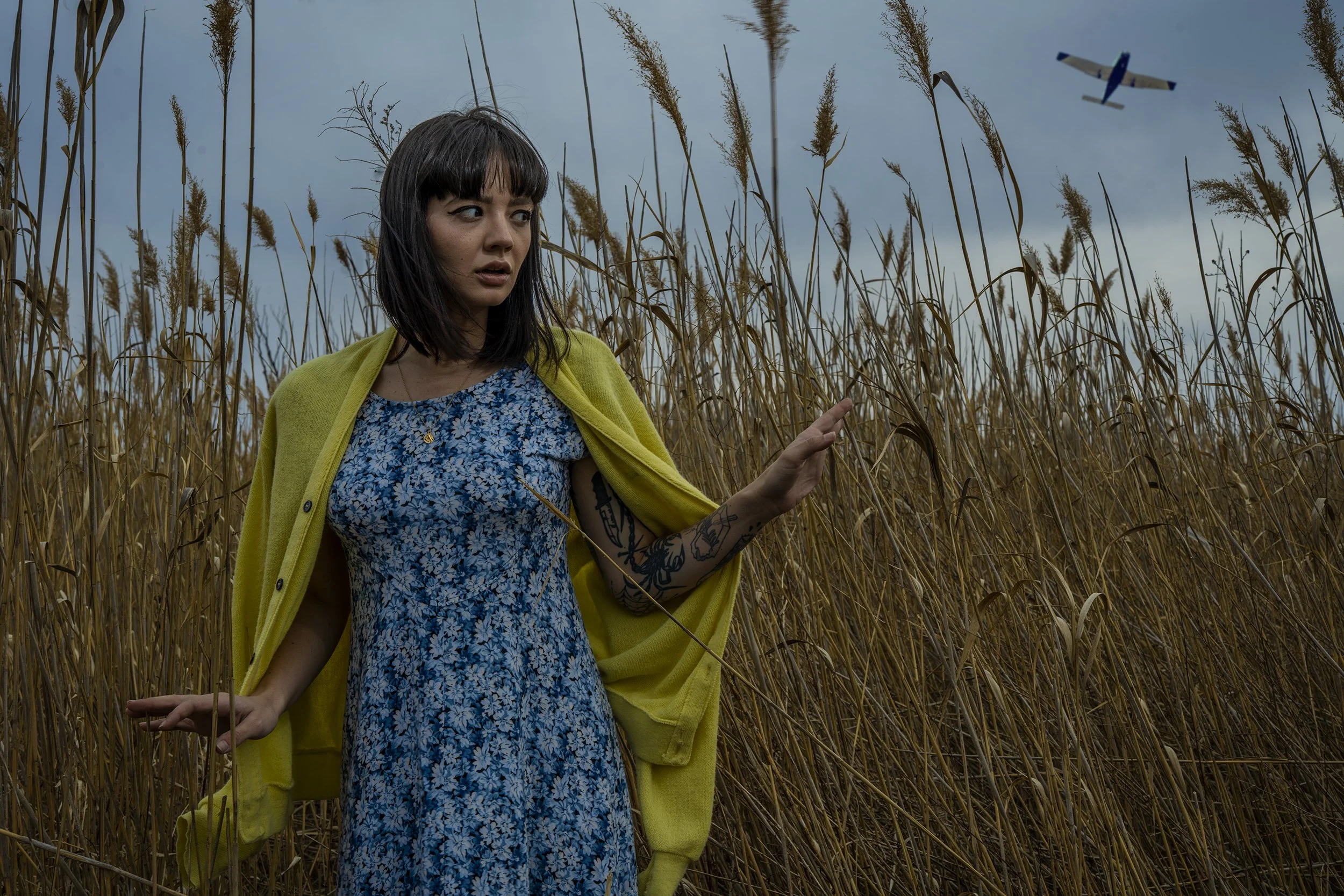 A woman with dark hair and tattoos on her arm standing in a wheat field, wearing a blue floral dress and a yellow cardigan, with an airplane flying in the overcast sky.