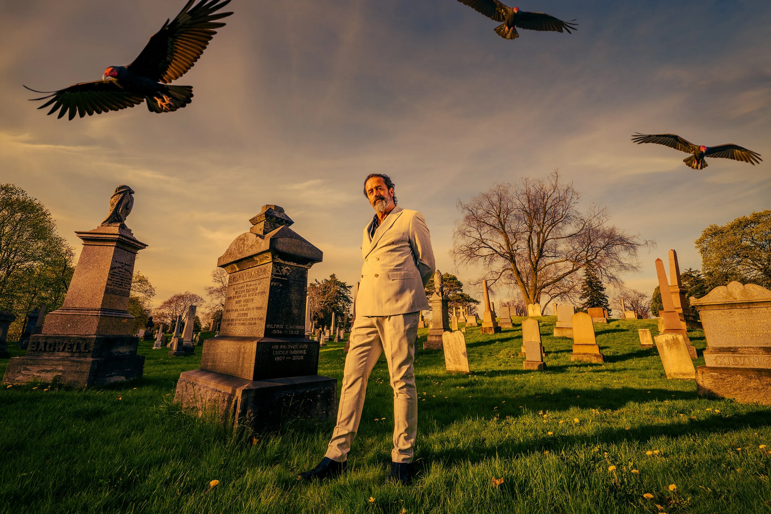 A man in a cream-colored suit standing in a cemetery with gravestones and trees, under a sky with flying birds at sunset.