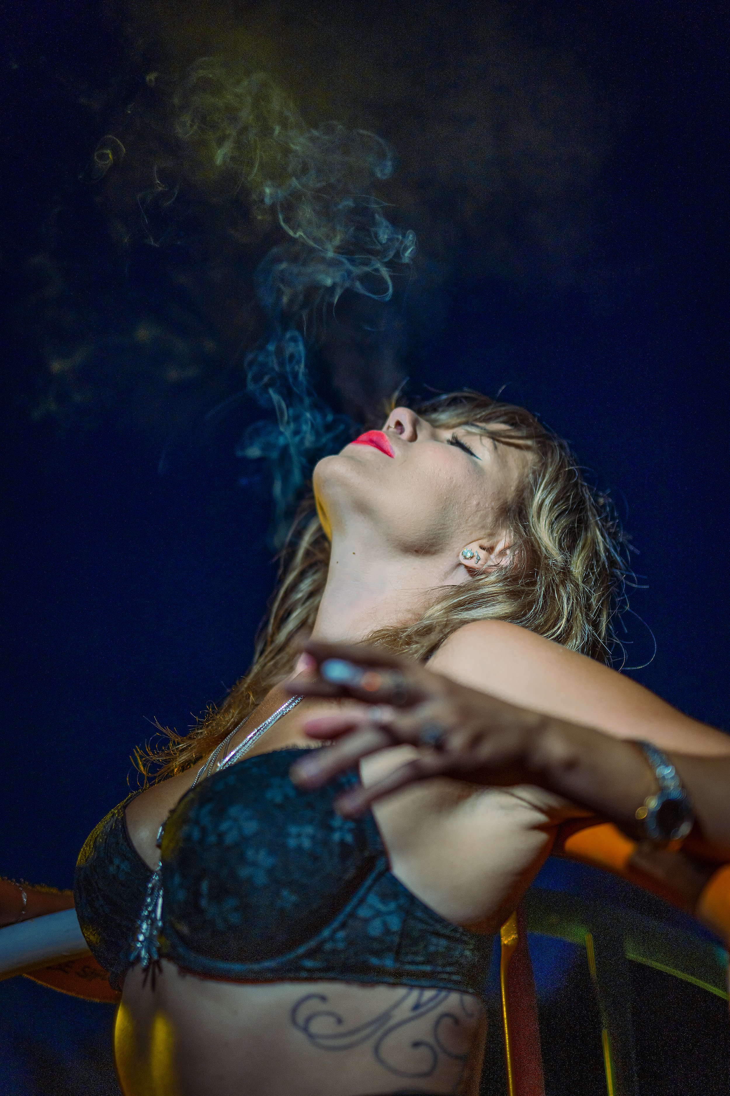 A woman with her head tilted back, eyes closed, exhaling smoke, wearing red lipstick, earrings, a black lace bra, and a tattoo on her side. The background is dark.