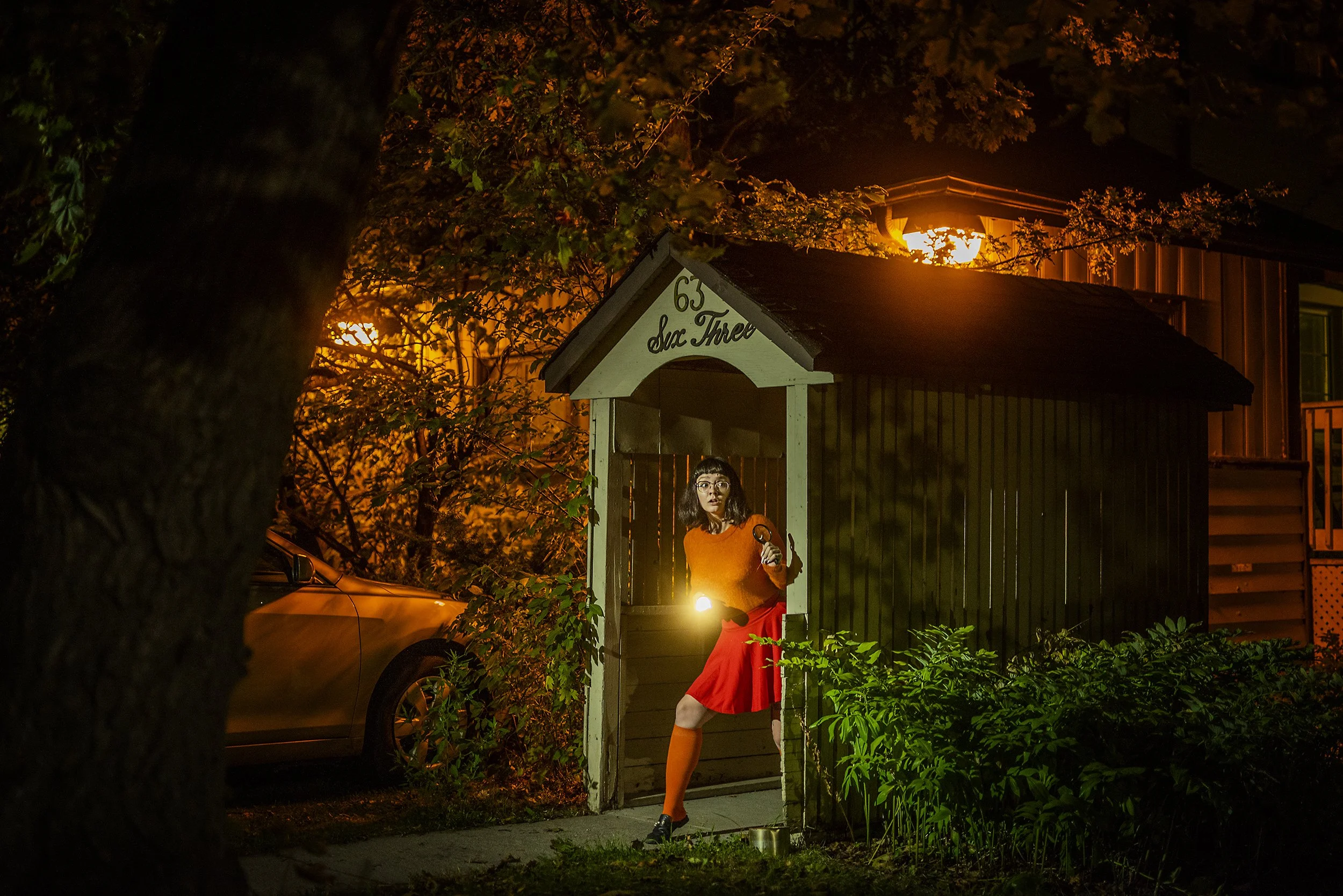 A woman standing in the doorway of a small shed at night, holding a flashlight in one hand and a cane in the other, illuminated by warm exterior lighting and surrounded by trees and bushes.