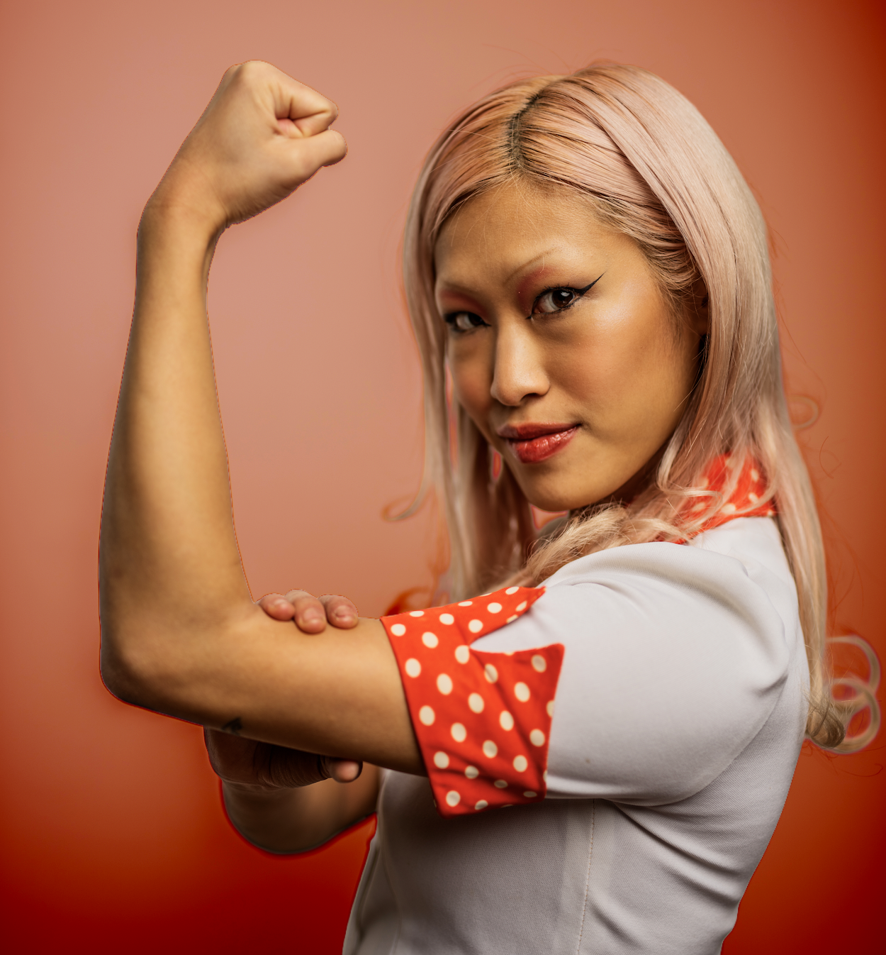 Woman with pink hair flexing her bicep, wearing a white shirt with red polka dot sleeves, on a pink background.