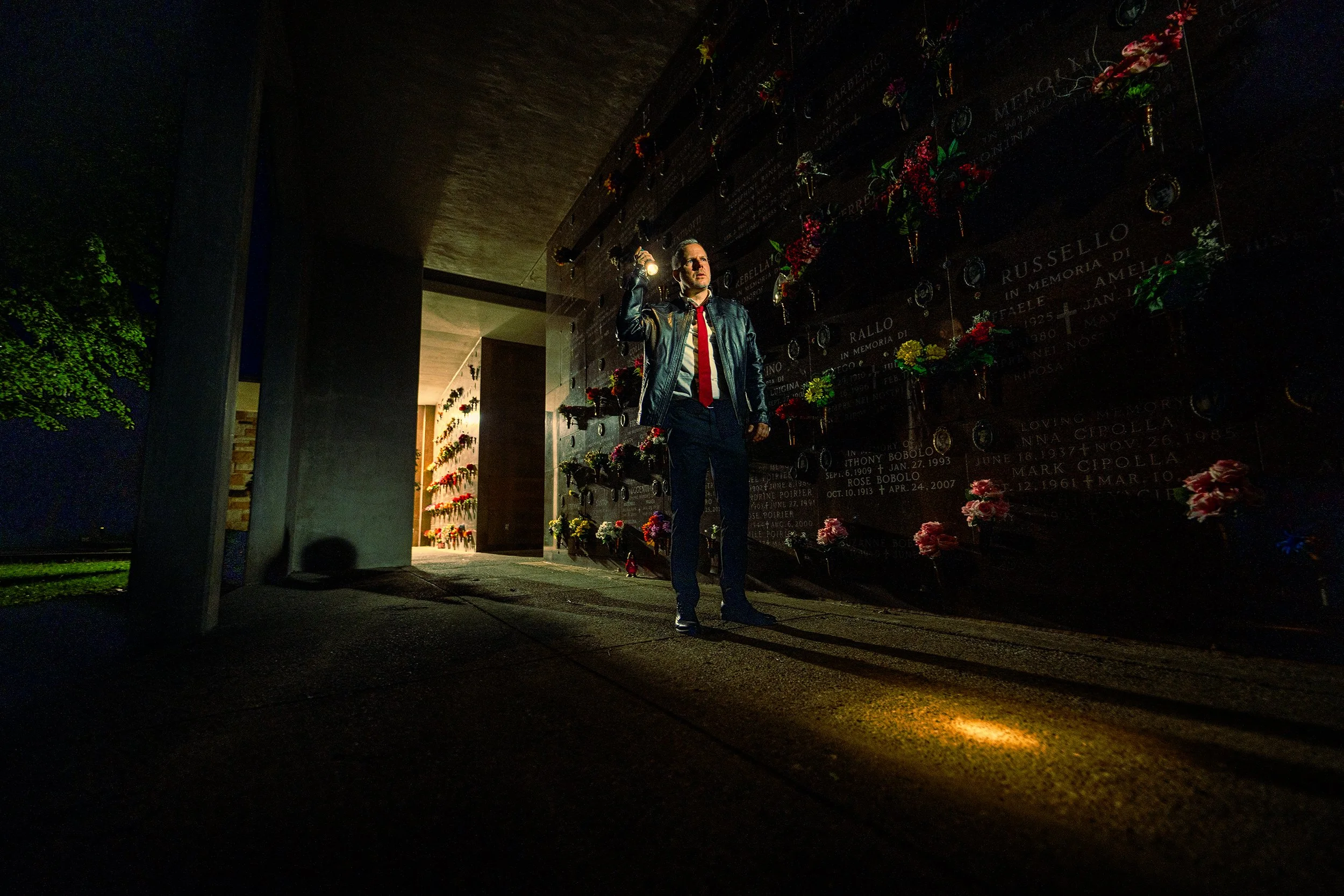 A man in a leather jacket and red tie holding a flashlight, standing in front of a memorial wall decorated with flowers and plaques, at night.
