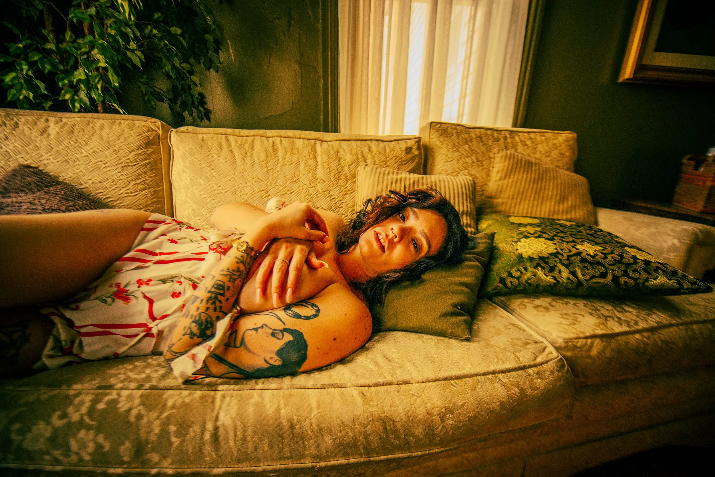 A woman with tattoos lying on a vintage sofa with cushions, in a warmly lit room with green plants and curtains.