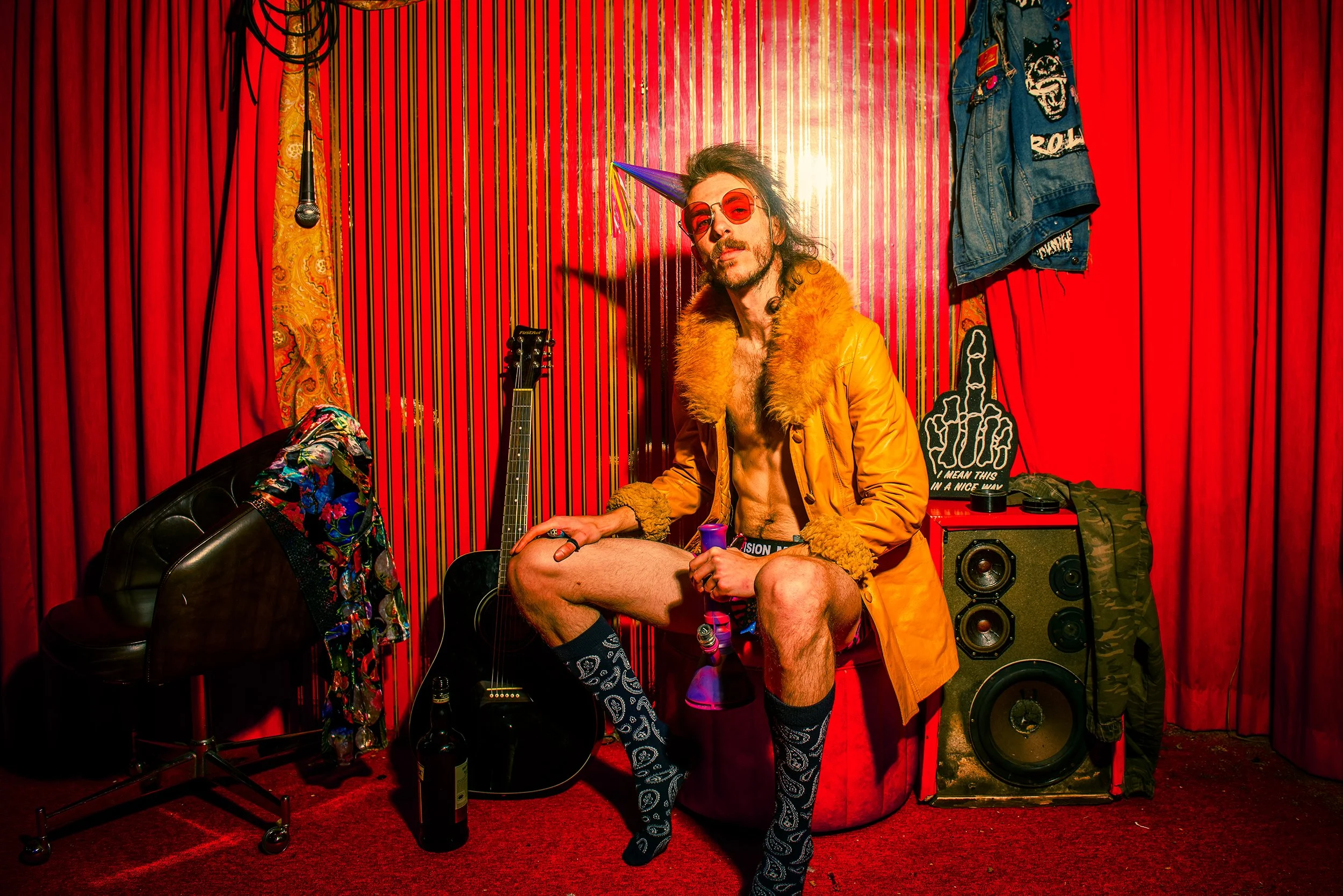 A man with long dark curly hair, wearing a purple party cone hat, red sunglasses, an orange fur-lined jacket, and patterned socks, poses in a vibrant room with red curtains. He is sitting on a black chair, holding a bottle, with a guitar, speaker, an