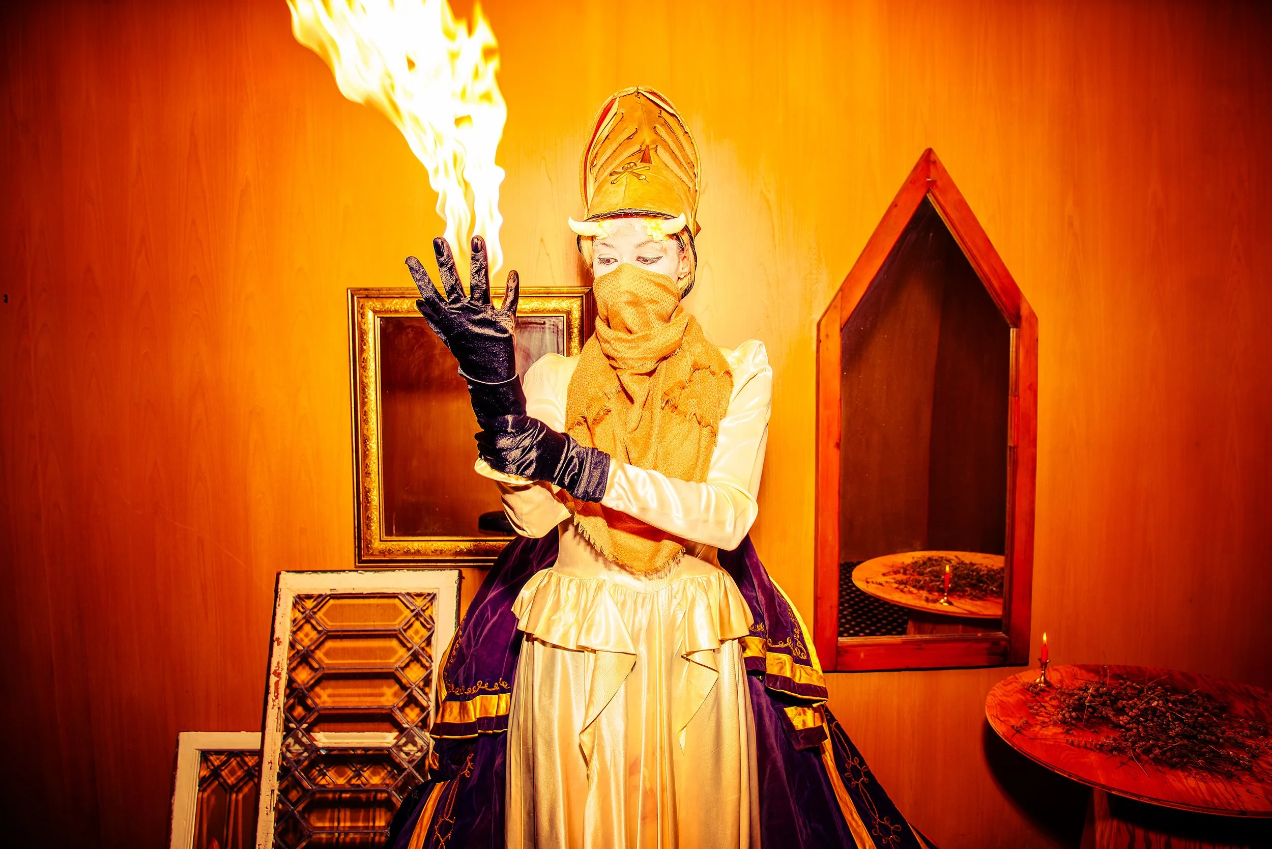 A person dressed in a costume with a large orange hat and mask, holding a flaming torch, standing in front of a wooden wall with frames and furniture.