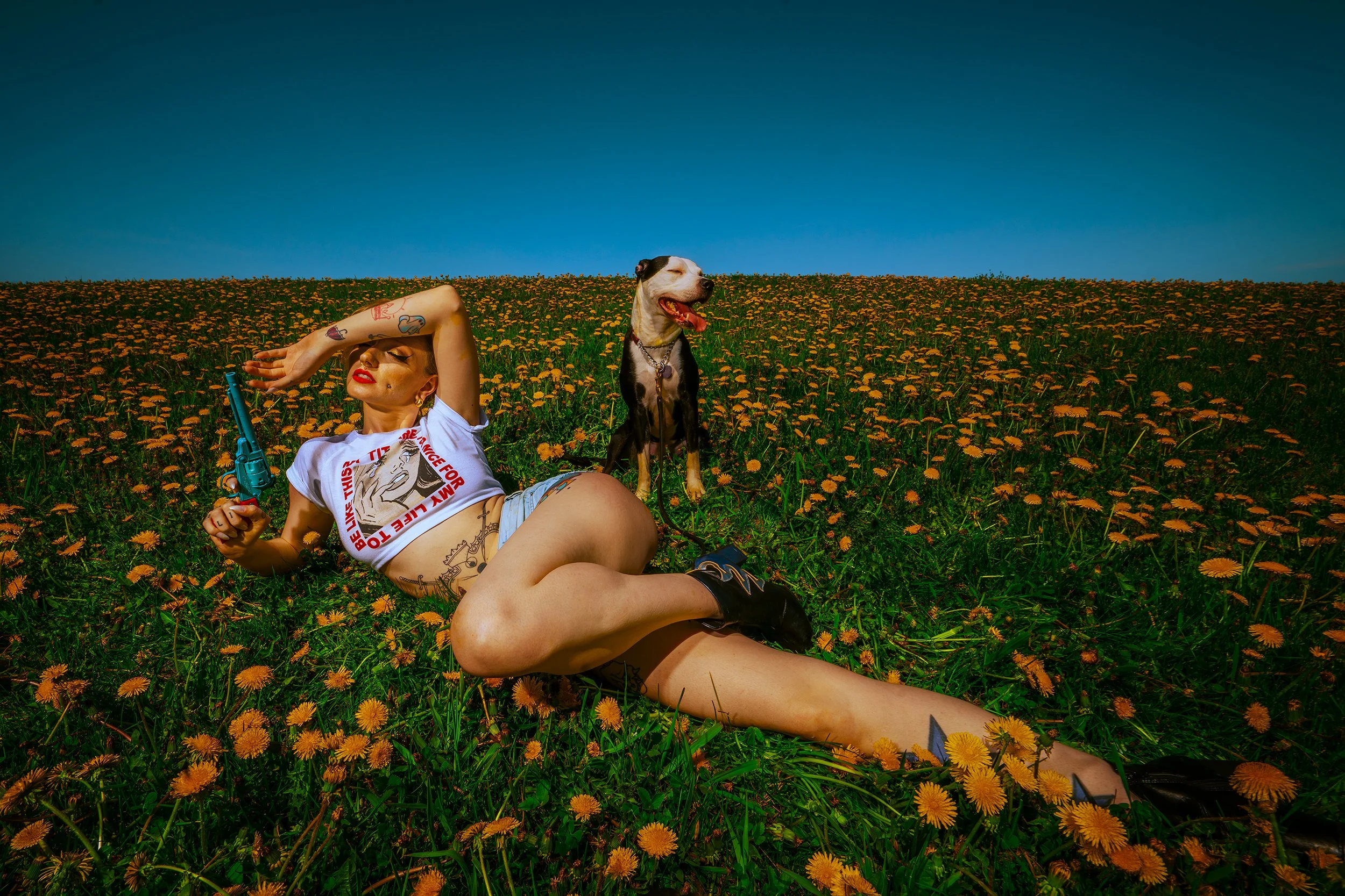 A woman with tattoos lying in a field of orange flowers with a dog sitting beside her. The woman holds a toy gun and is wearing a white graphic t-shirt, shorts, and black shoes. The sky is clear and blue.