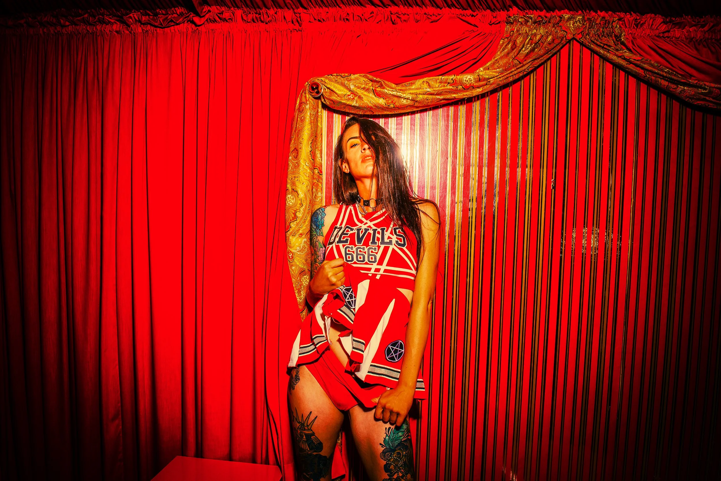 A woman with black hair and tattoos wearing a red basketball jersey, standing in front of red curtains and red wall decor, with dramatic lighting highlighting her face.