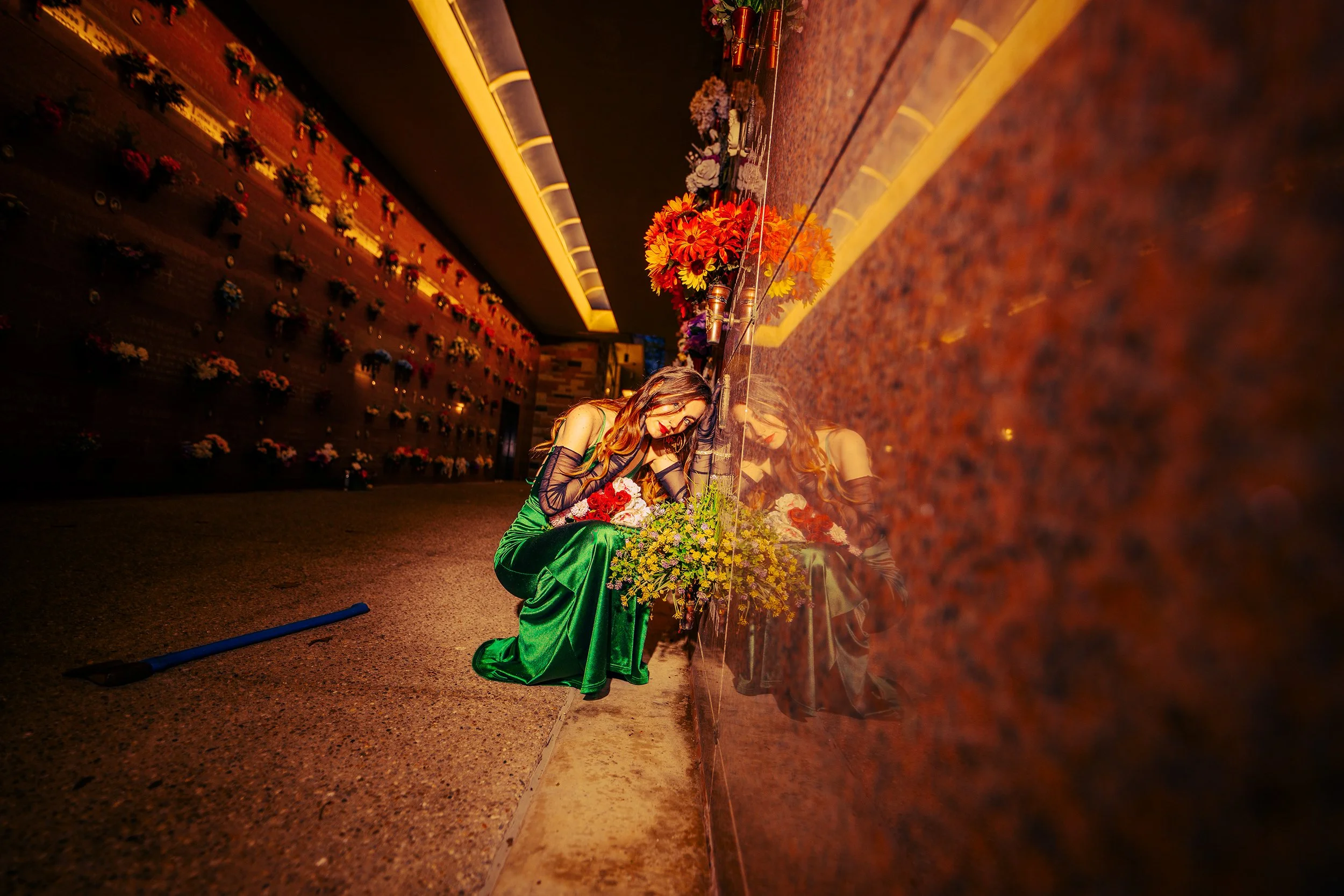 A woman in a green dress with black lace gloves kneels beside a floral arrangement, looking down, in front of a reflective polished surface that shows her reflection. The background is decorated with various flowers on a wall, illuminated by warm yel