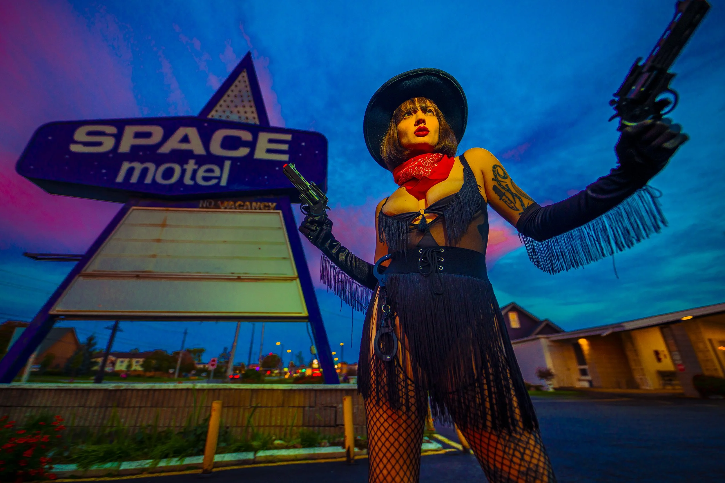A woman dressed in a black fringe dress, fishnet stockings, and a wide-brimmed hat, standing outside the Space Motel sign at dusk, holding a toy gun in each hand.