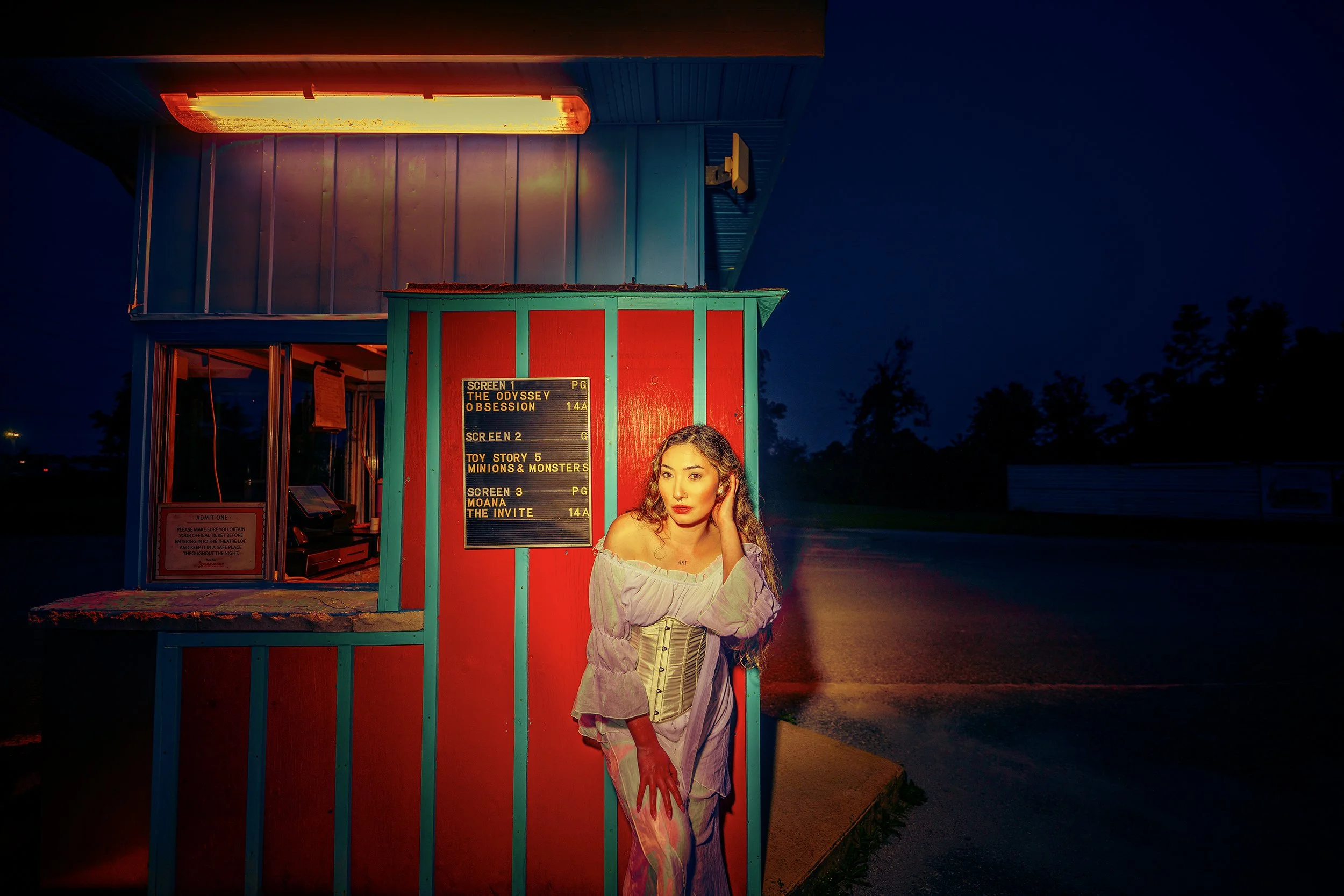 A young woman with long, wavy hair standing in front of a colorful movie theater ticket booth at night. She is wearing a light-colored off-shoulder top and pants, with one hand resting on her temple. The theater marquee displays upcoming movies on a 
