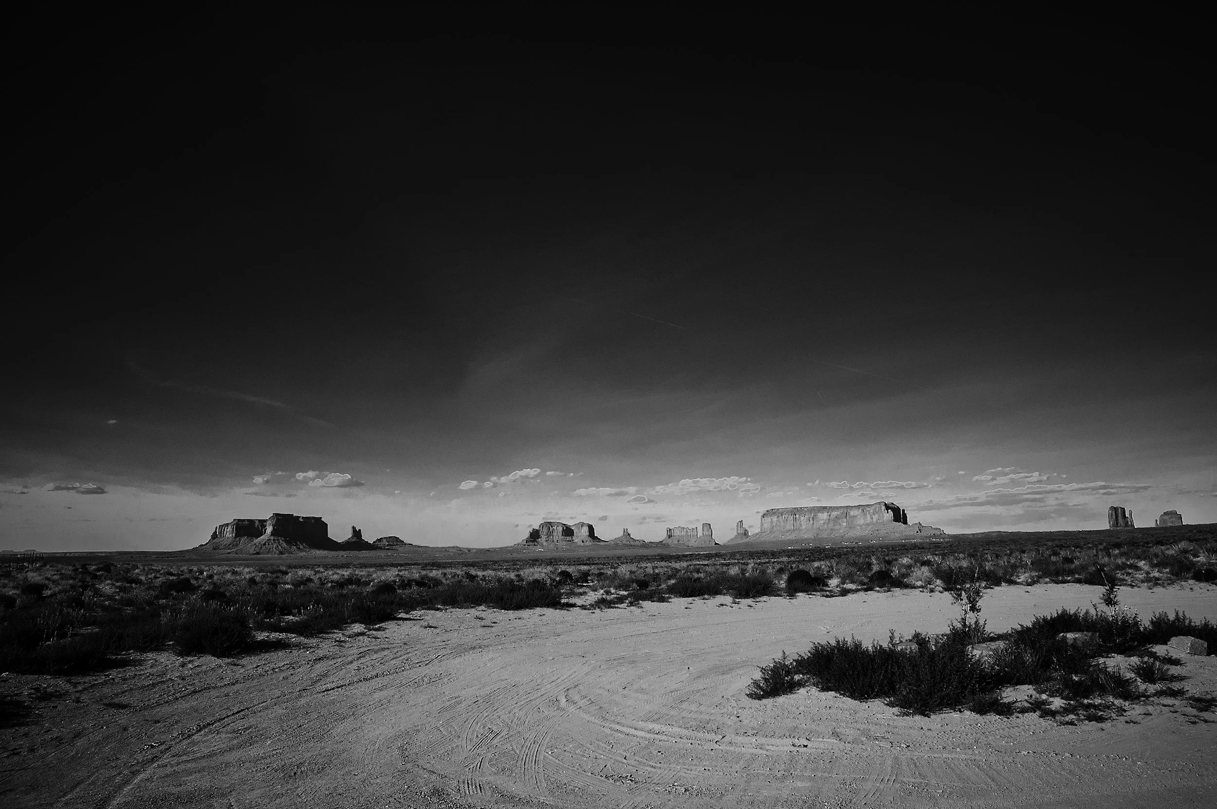 Black and white photo of desert landscape with rock formations, sparse vegetation, and a wide sky with clouds.