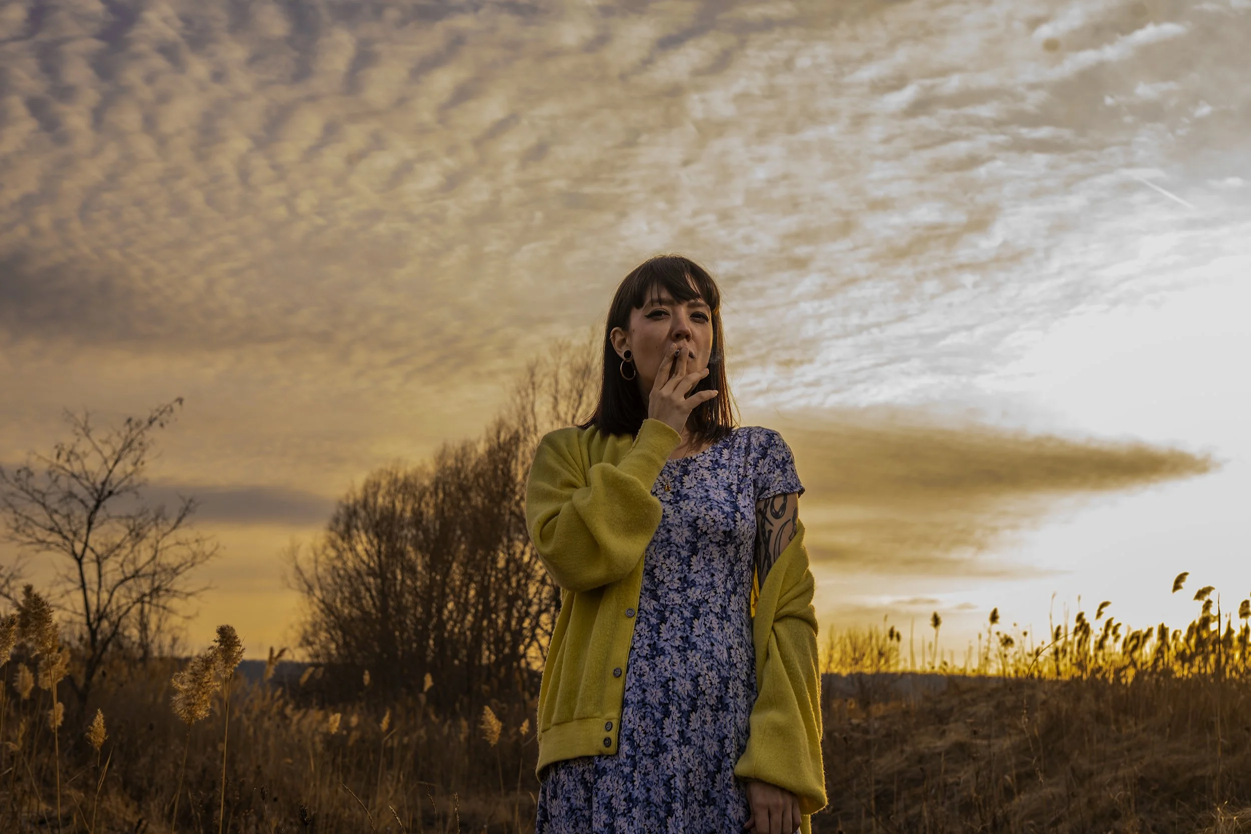 A woman in a floral dress and yellow cardigan standing outdoors during sunset, with trees and tall grass in the background.