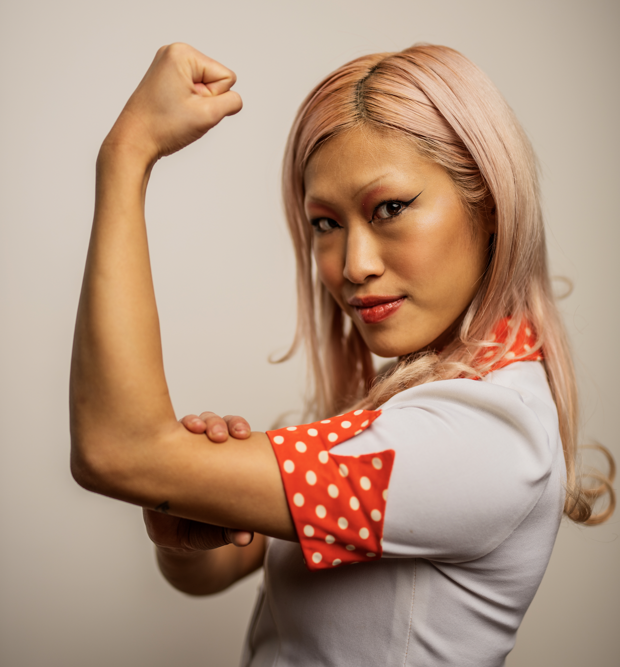 Woman with pink hair and makeup flexes her arm in a strength pose, wearing a white shirt with red polka-dot accents.