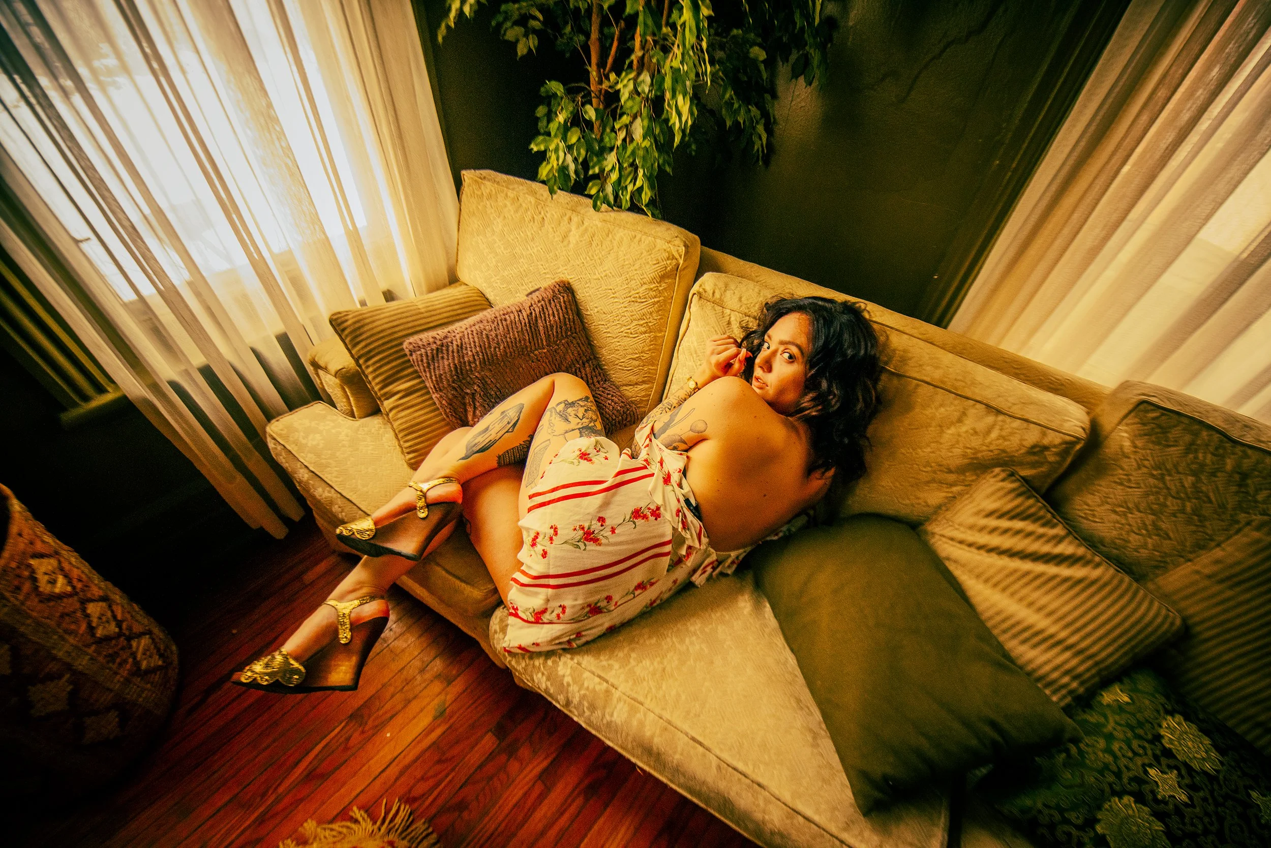 A woman with dark curly hair and tattoos on her arm and leg, wearing a white dress with red stripes and floral pattern, sitting on a yellow sofa in a room with dark walls, curtains, and a large plant, holding a phone to her ear and looking at the cam