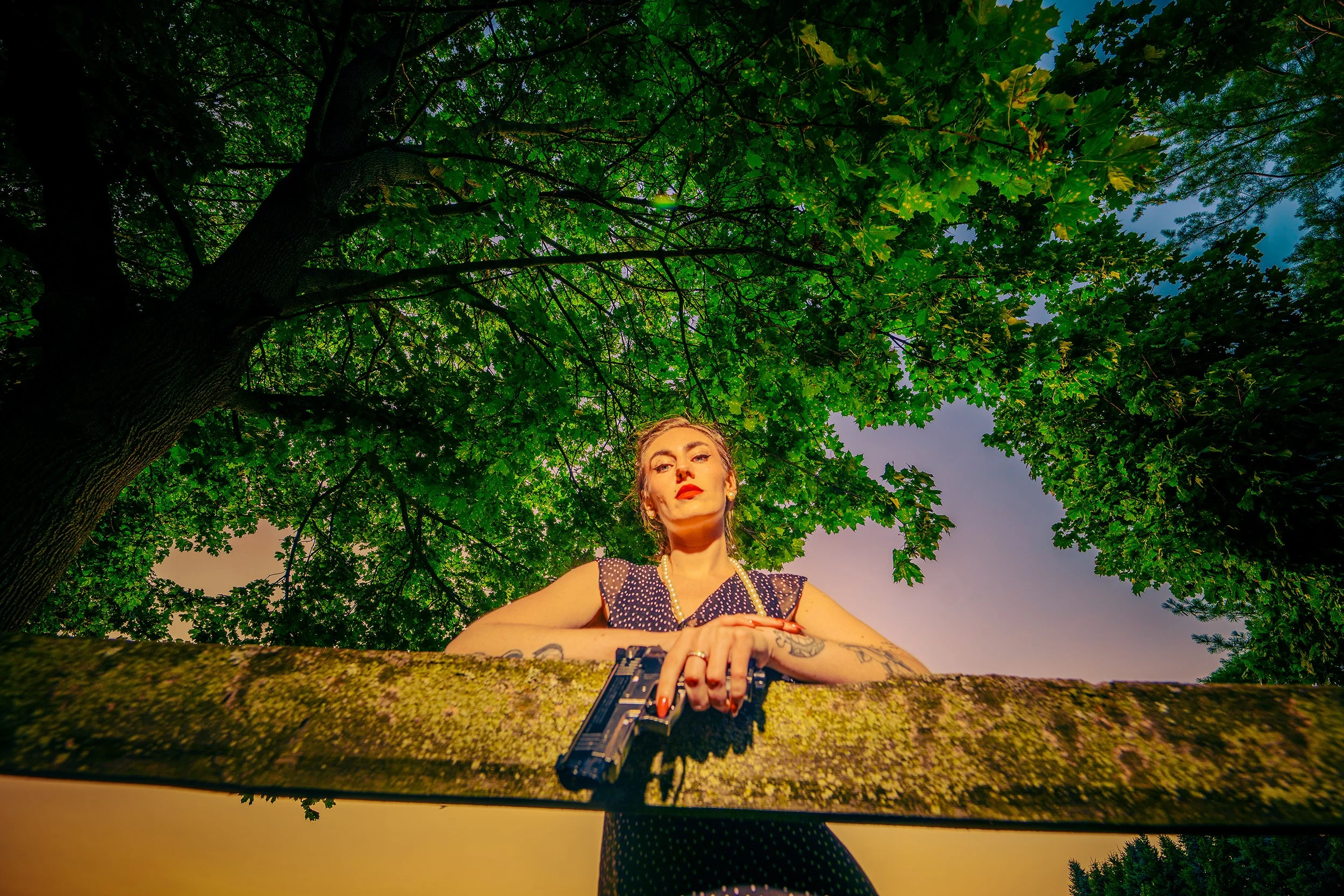 A young woman with tattoos and vintage-inspired makeup stands outdoors beneath a large leafy tree, holding a handgun pointed downward at waist level, with the sky and green foliage in the background.