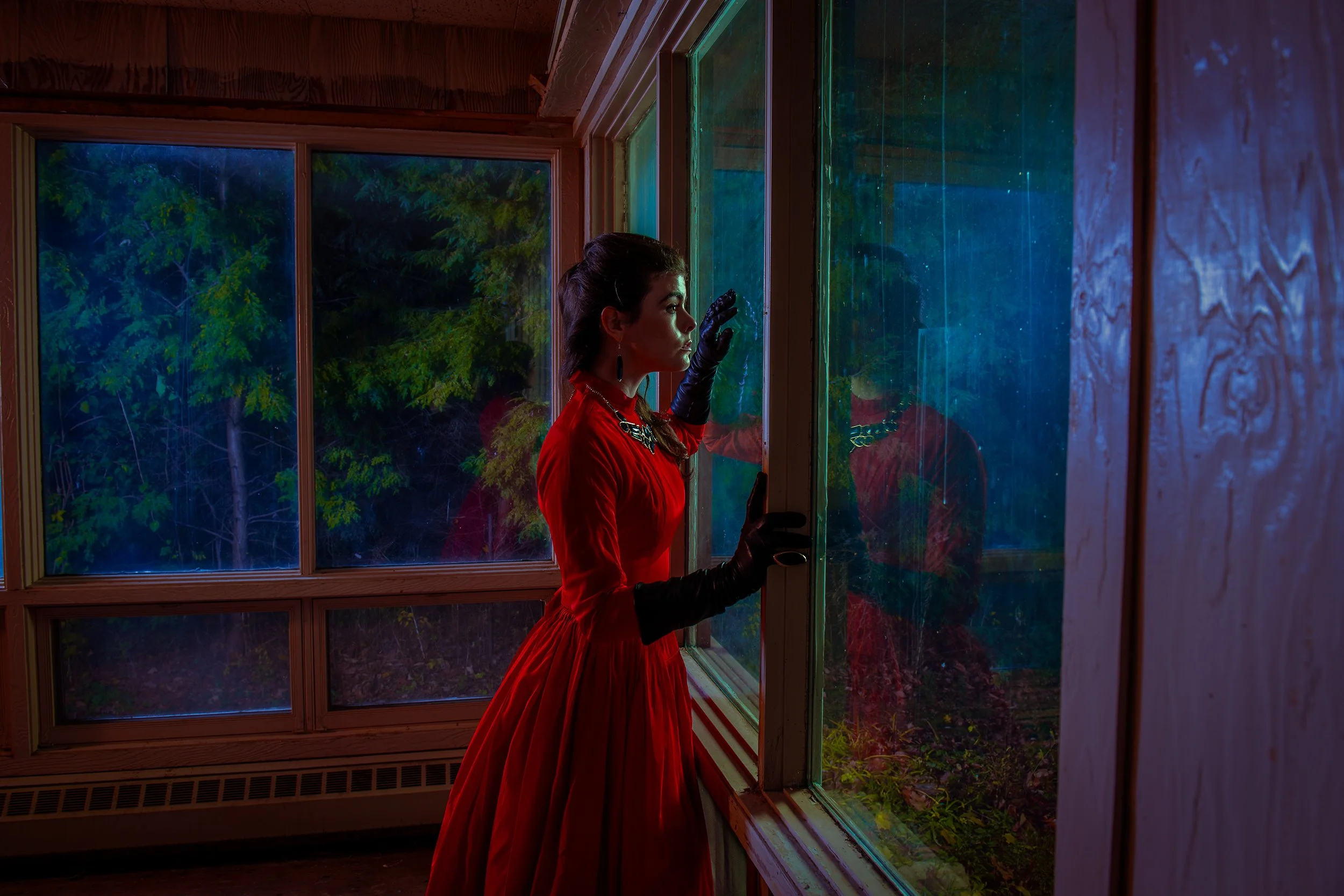 A woman in a red dress with black gloves and earrings, looking out a large window of a wooden building at nighttime, with trees visible outside. The window has multiple panes, some with colored reflections.