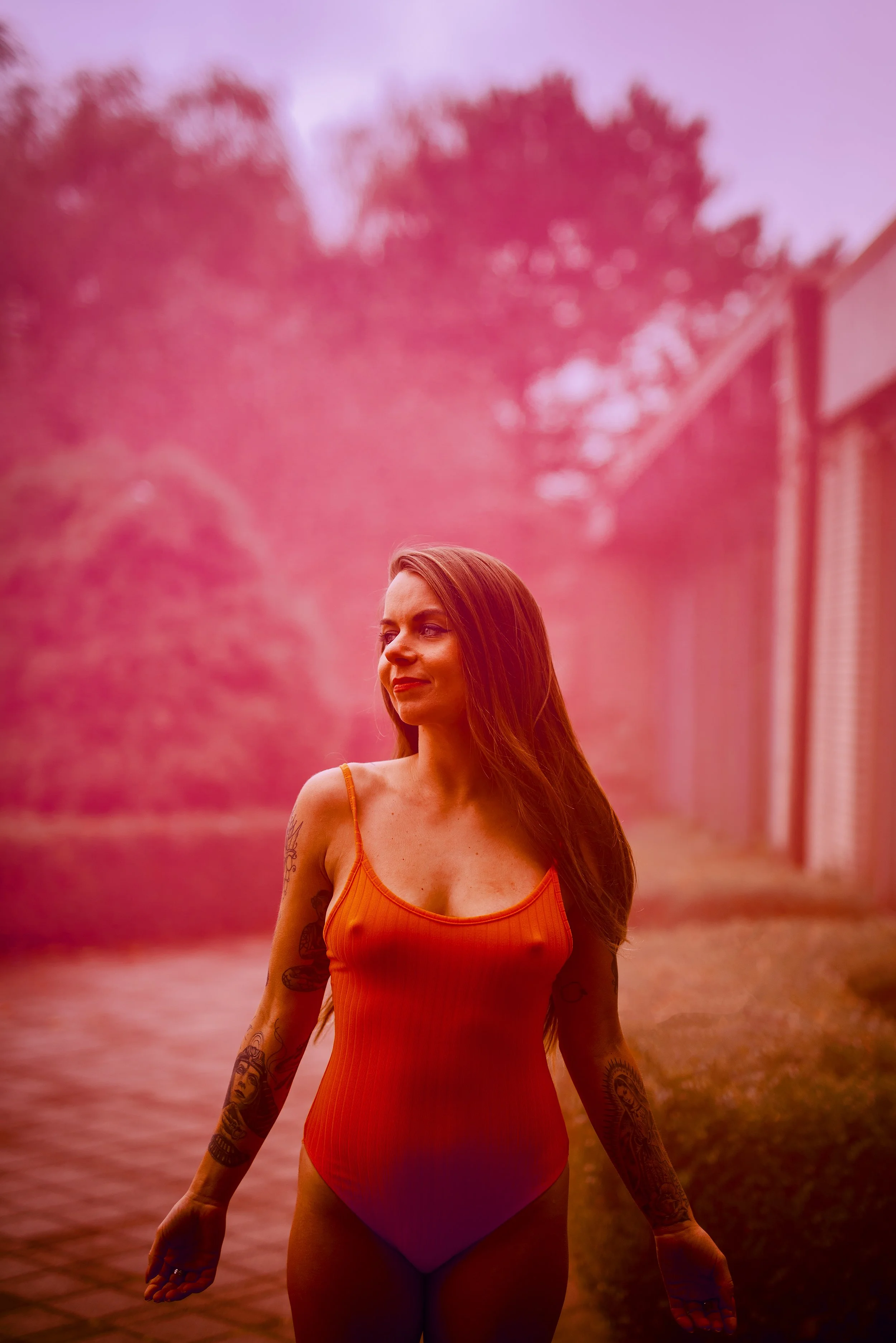 A woman with long reddish hair and tattoos on her arms standing outdoors during sunset, wearing an orange tank top and purple shorts, with pink and purple foliage in the background.