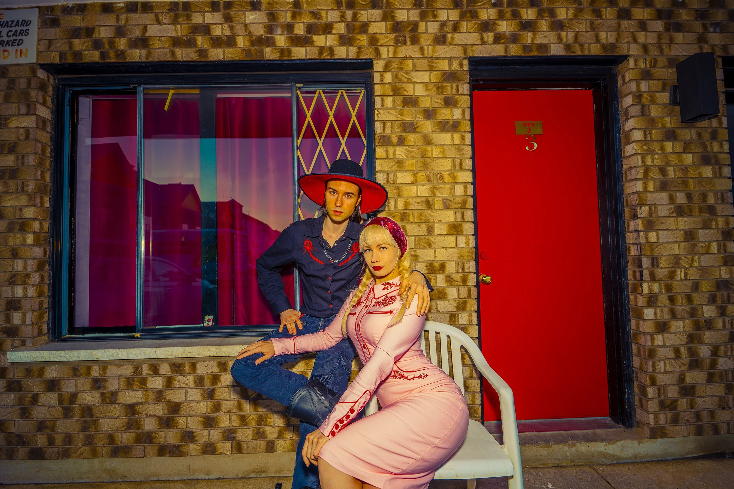 A young man with long hair wearing a black hat and a dark shirt stands beside a young woman with blonde hair in a pink vintage-style dress with red embroidery. They are posed in front of a brick building with a large window and a bold red door. The w