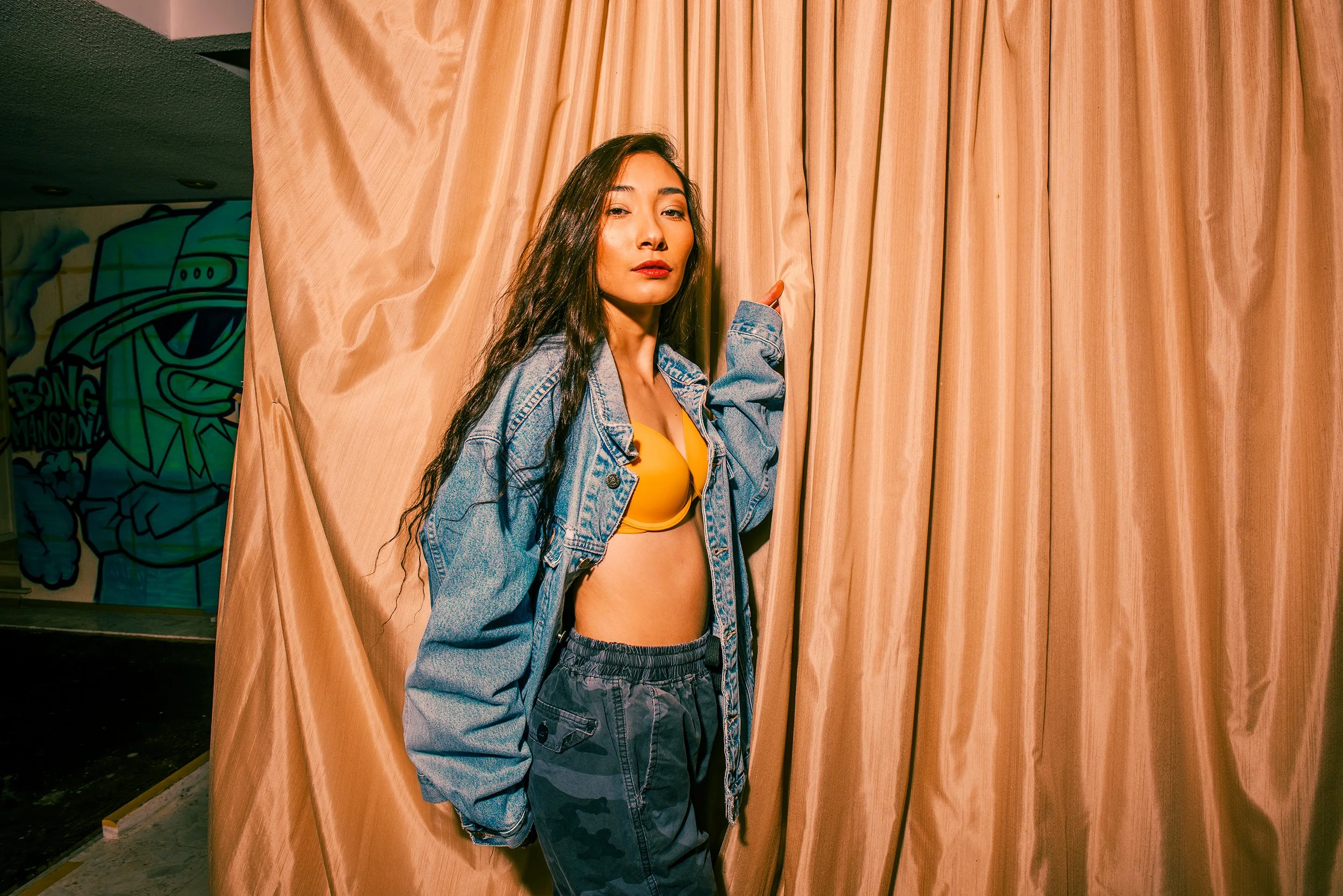 A young woman with long dark hair wearing a yellow top and a denim jacket, standing in front of beige curtains, holding one side of the curtain with her right hand.