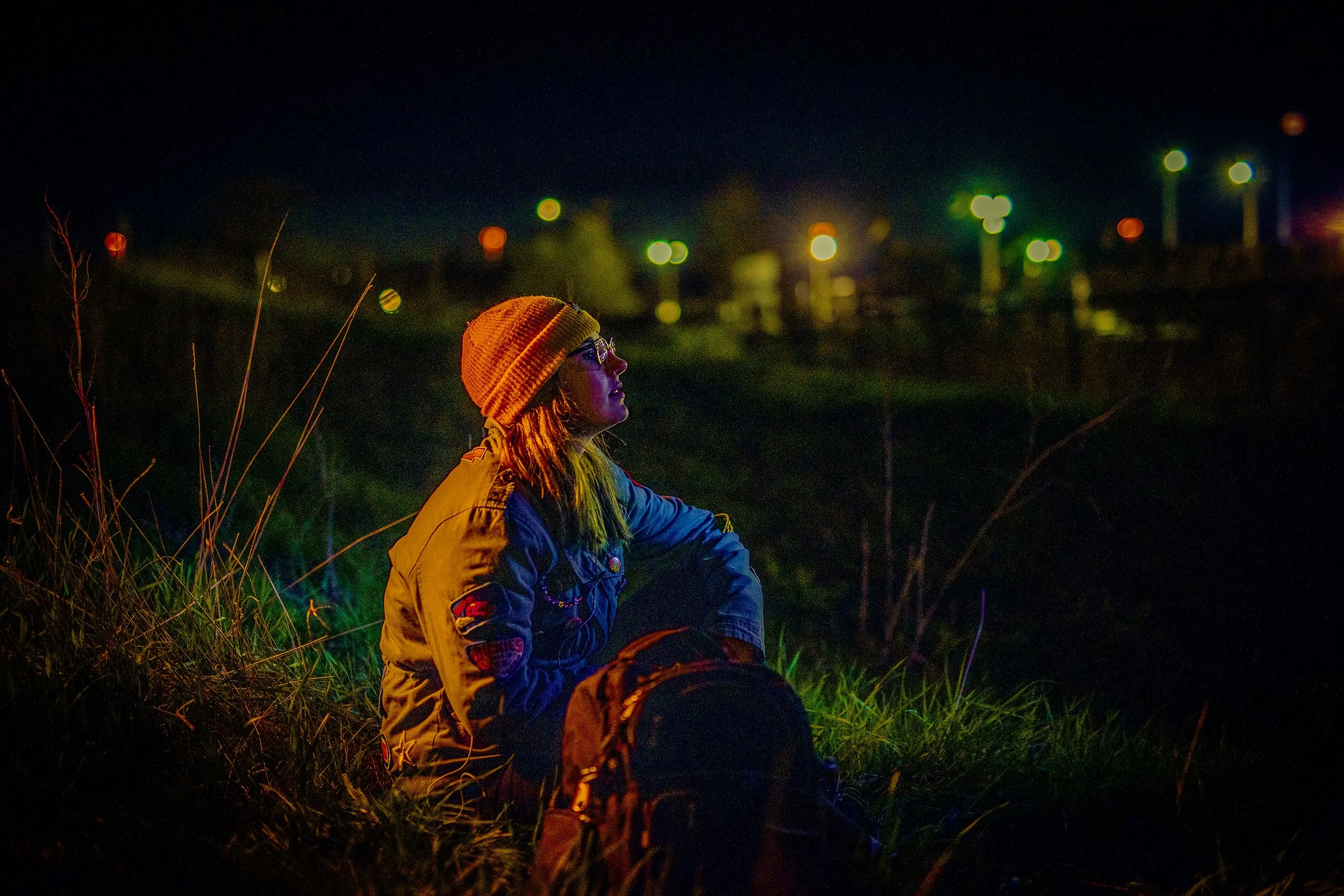 A person with long hair wearing a yellow beanie, glasses, and a colorful jacket is sitting on grass at night, looking thoughtfully into the distance. The background features blurred colorful lights against a dark sky.