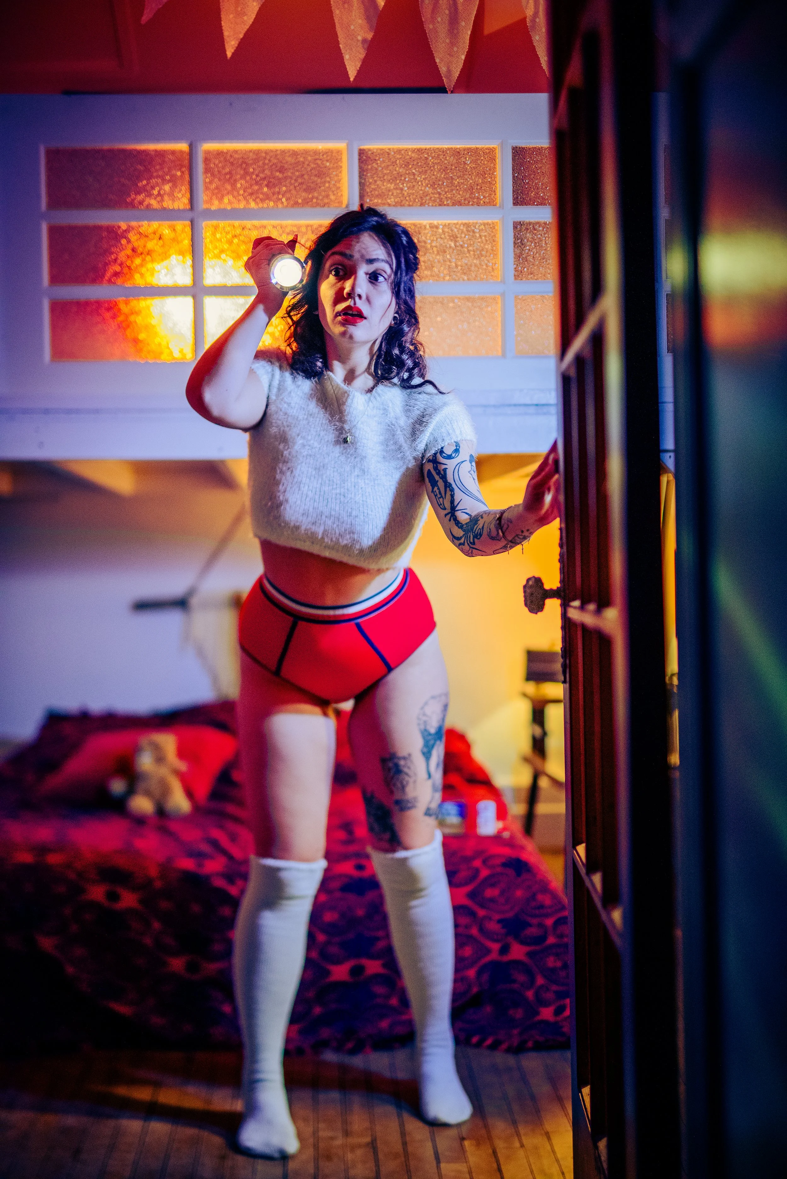 A woman with dark, curly hair holding a flashlight in her right hand, standing in a room with a red-themed bed and a colorful stained-glass window. She is wearing a white cropped sweater, red underwear, thigh-high white socks, and has tattoos on her 