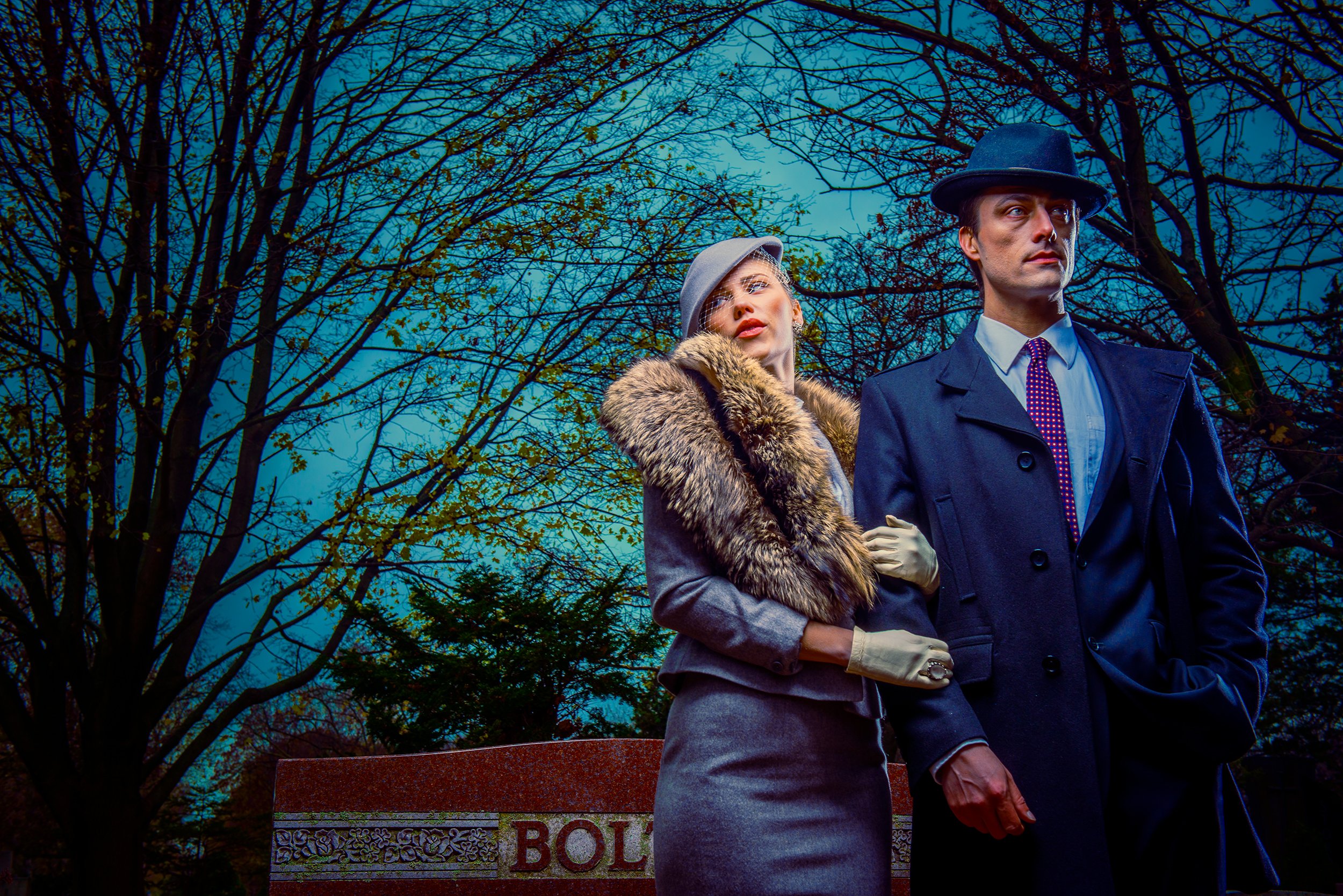 A man and woman dressed in vintage clothing standing outdoors near a bench with a sign that reads 'Pol' visible. The woman is wearing a gray hat, fur collar coat, and white gloves, while the man is in a dark coat, light blue shirt, red polka dot tie,